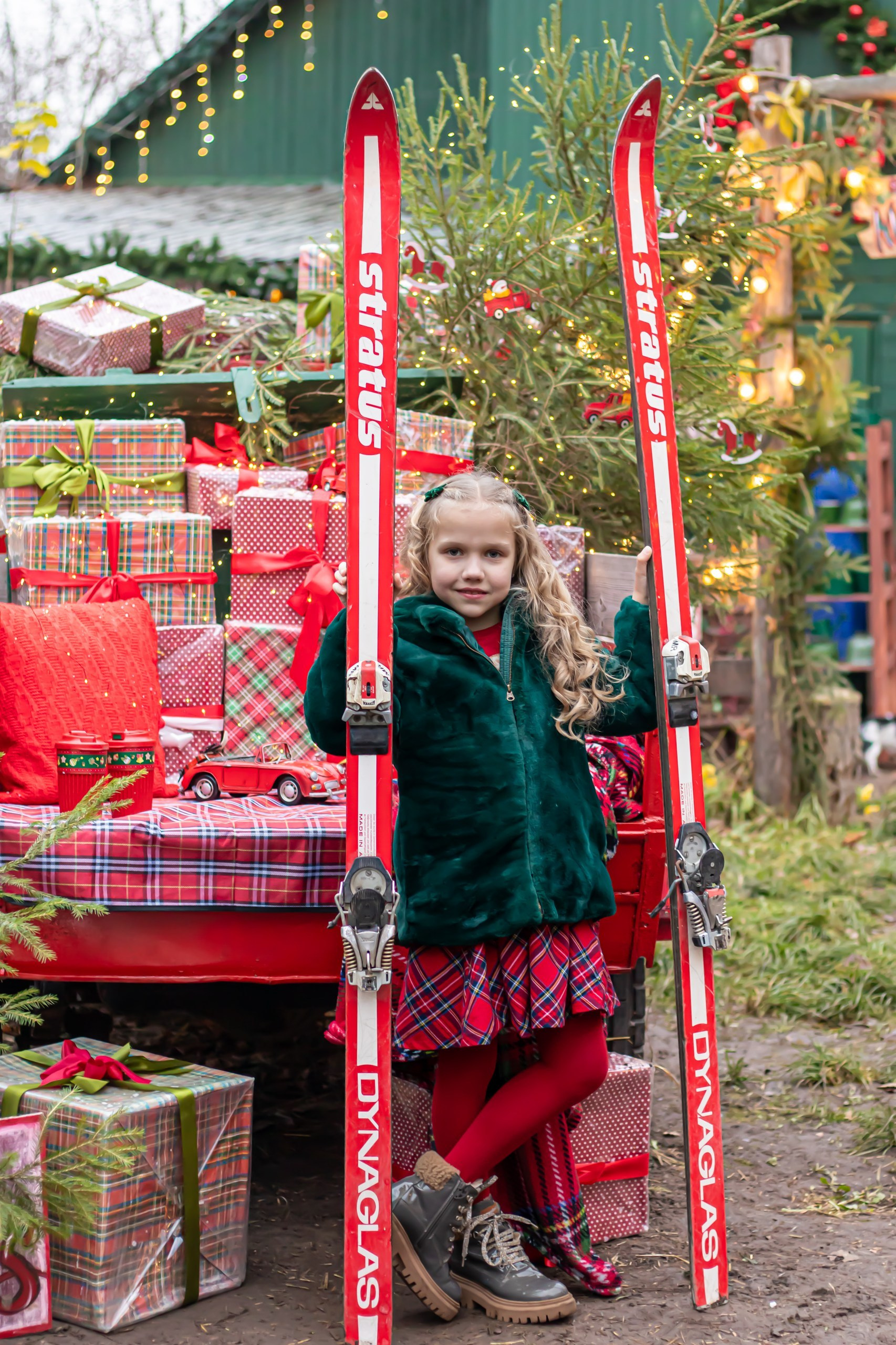 Девочка стоит на фоне рождественской площадки с огоньками, зелёная шубка и клетчатая юбка, фотограф Карасёва Елена