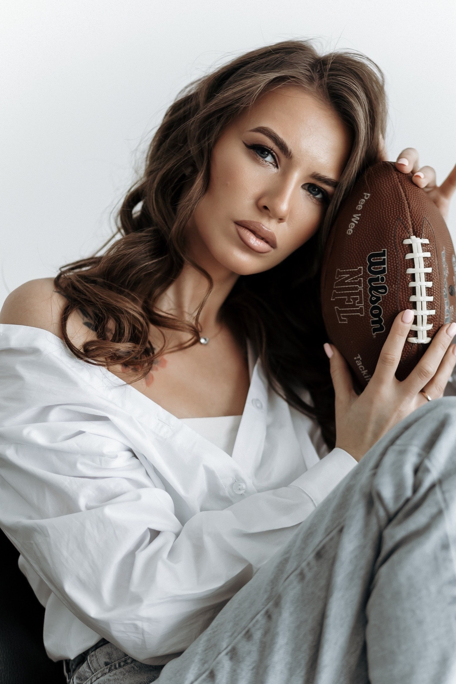 Девушка в белой рубашке позирует с американским футбольным мячом. Фон нейтральный.