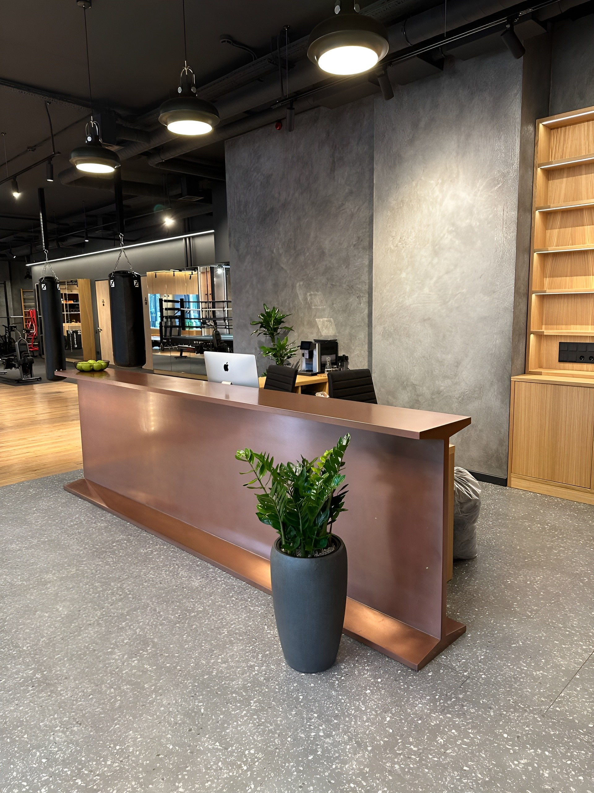 Этот проект для зала единоборств стал настоящим испытанием на прочность — как конструкции, так и нашего мастерства. Задача звучала амбициозно: создать стойку ресепшн в форме двутавровой балки, которая должна была не просто встречать посетителей, но и транслировать характер клуба — силу, надежность, индустриальную эстетику. Конструктивный вызов: Форма двутавра сама по себе сложна в изготовлении из листовых материалов. Необходимо было спроектировать каркас из МДФ и фанеры, который выдержит эксплуатационные нагрузки, сохранит геометрию и при этом будет выглядеть как настоящая металлическая балка. Технология отделки: Главная сложность заключалась в создании эффекта настоящей меди. Мы использовали металлизированную краску с реальными частицами металла в составе. Процесс был многоэтапным и требовал ювелирной точности: Не менее 5 слоев грунта, каждый из которых тщательно шлифовался3 слоя металлизированной эмали с промежуточной шлифовкой. Финальная полировка до состояния, неотличимого от натуральной меди. Любая ошибка на любом этапе означала бы перекраску всего изделия с нуля. Монтаж: Установка потребовала профессиональных такелажных работ и абсолютной аккуратности. Малейший скол многослойного покрытия обнулил бы недели работы. Каждое движение было выверено, каждая точка контакта защищена. Результат — монументальная стойка ресепшн, которая встречает бойцов как символ силы и несгибаемости, выполненная с точностью ювелирного изделия.