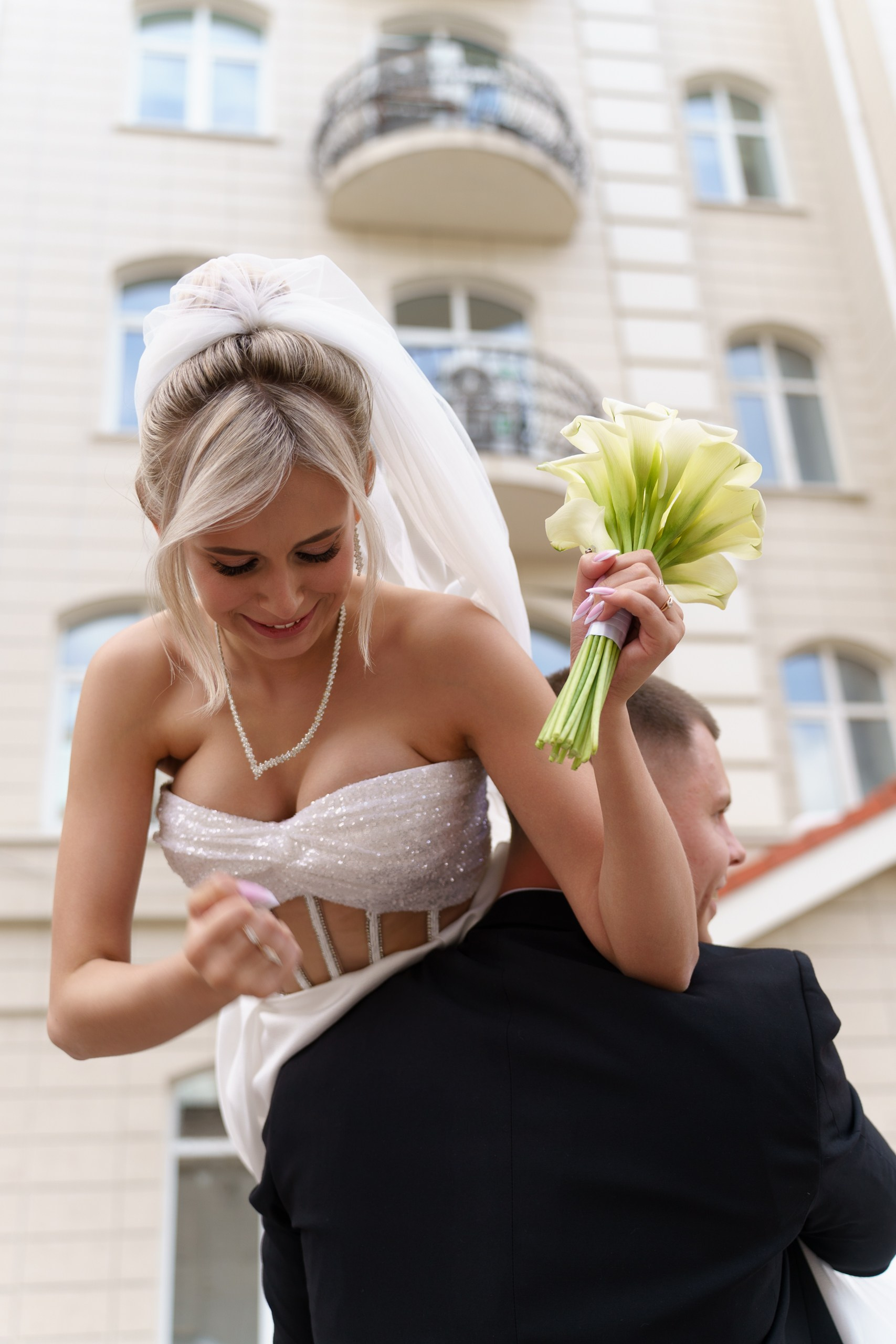 Жених несёт свою невесту во время свадебной фотосессии. Она одета в блестящее белое платье, её волосы уложены в элегантную причёску, а в руке — букет светло-зелёных цветов | экспрессивные свадебные фотографии Екатеринбург