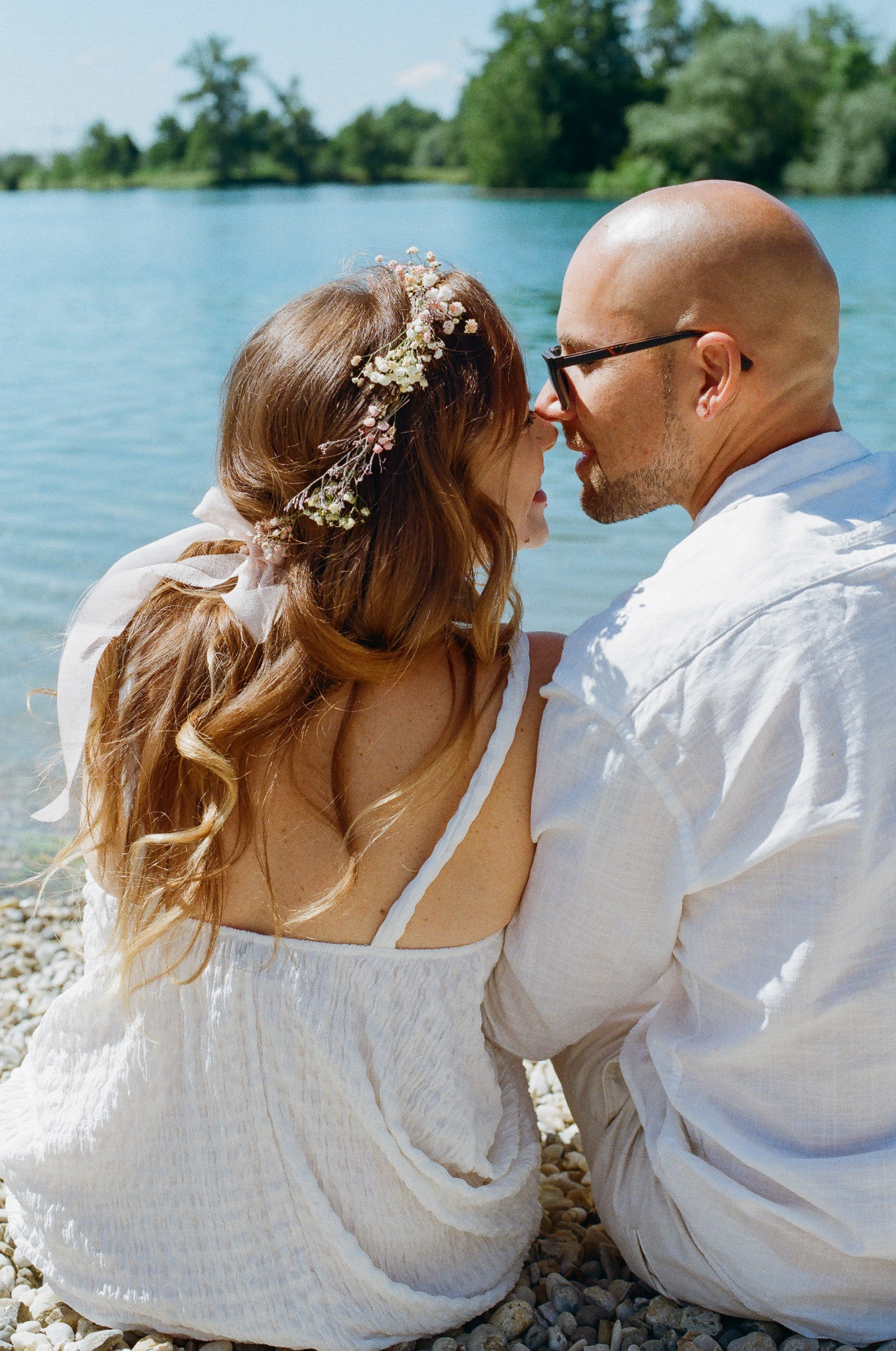 A couple kissing each other on the riverbank wearing white clothes and flower wreath