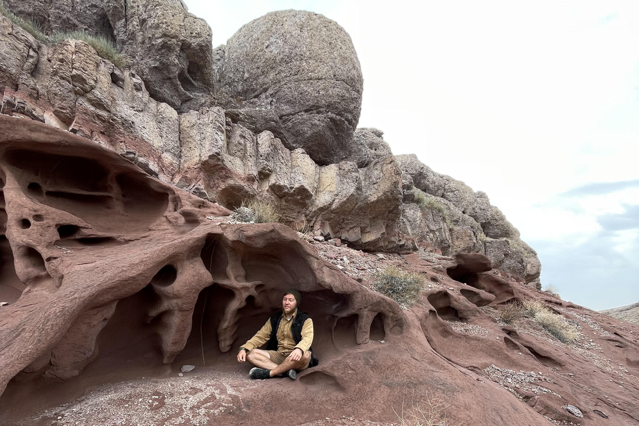 Человек позирует на фоне бирюзового озера Иссык в районе Алматы и гор. Он стоит на большом камне, одет в ярко-зеленую толстовку, черные штаны и светлую обувь, а также оранжевую панаму. В руках держит очки. За ним виднеются деревья, лесистые склоны и горные вершины под облачным небом. Атмосфера спокойная и природная.
