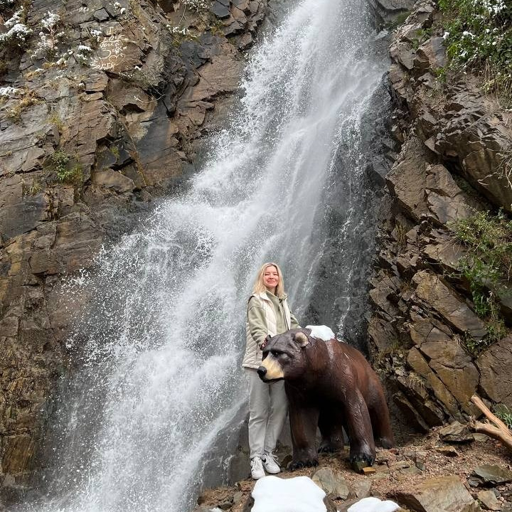 Женщина стоит рядом со скульптурой медведя на фоне красивого Медвежьего водопада в районе Алматы, спадающего по скалистым утёсам. На вершине скал и кустарниках виден свежевыпавший снег, что подчёркивает прохладную атмосферу. Каменистая местность у подножия водопада добавляет природной аутентичности сцене.