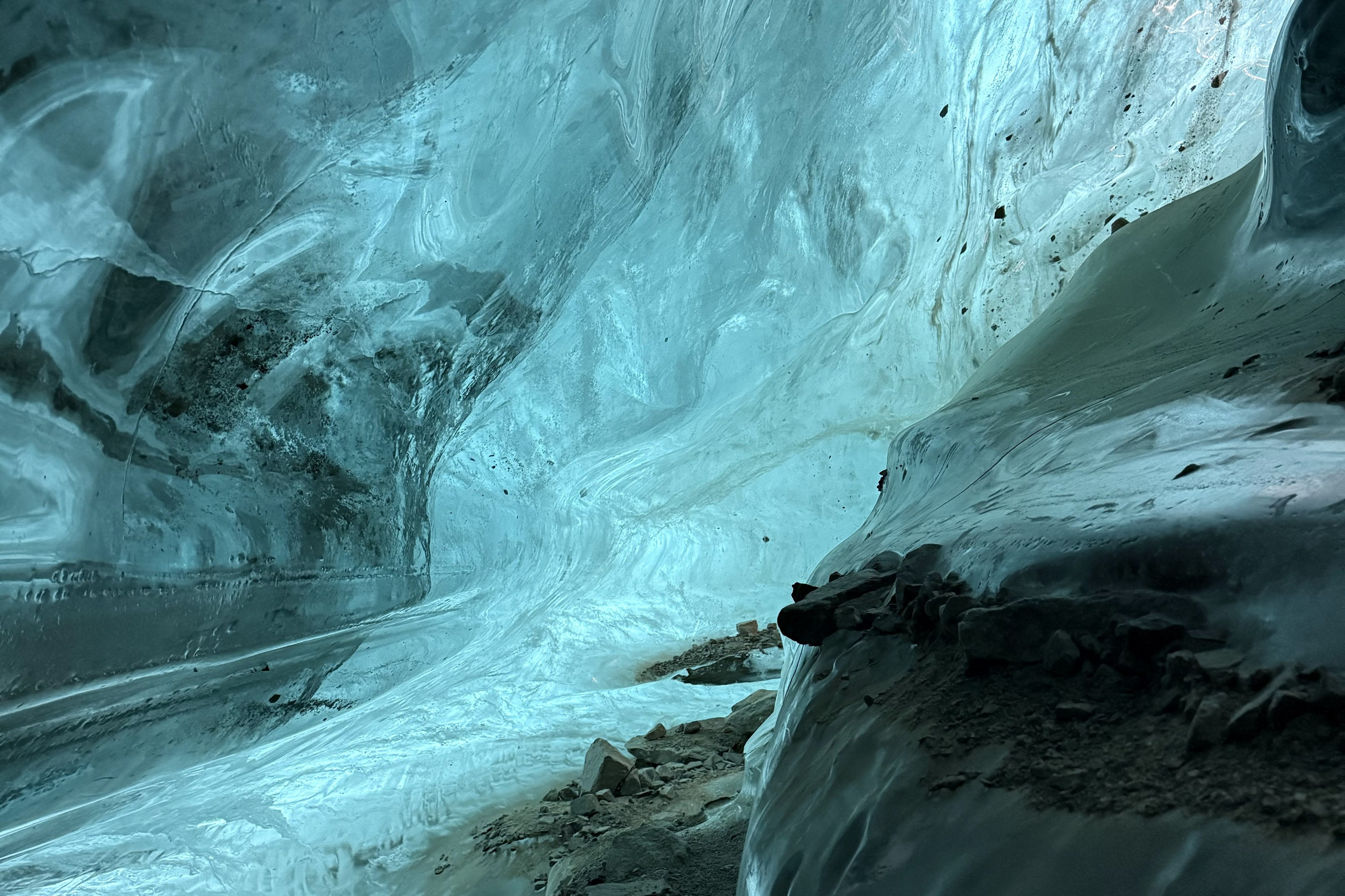 Человек позирует на фоне бирюзового озера Иссык в районе Алматы и гор. Он стоит на большом камне, одет в ярко-зеленую толстовку, черные штаны и светлую обувь, а также оранжевую панаму. В руках держит очки. За ним виднеются деревья, лесистые склоны и горные вершины под облачным небом. Атмосфера спокойная и природная.