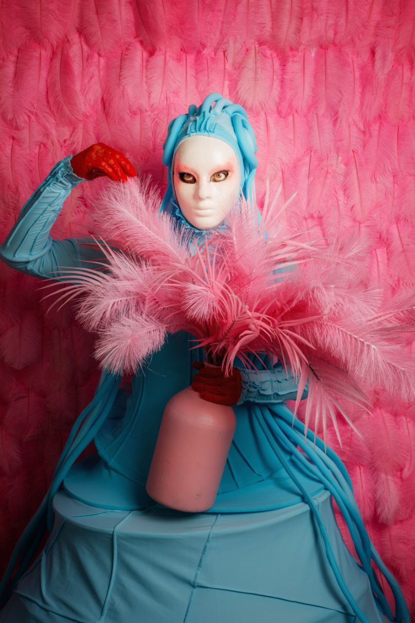 Мим в голубом костюме держит вазу с искусственными цветами.