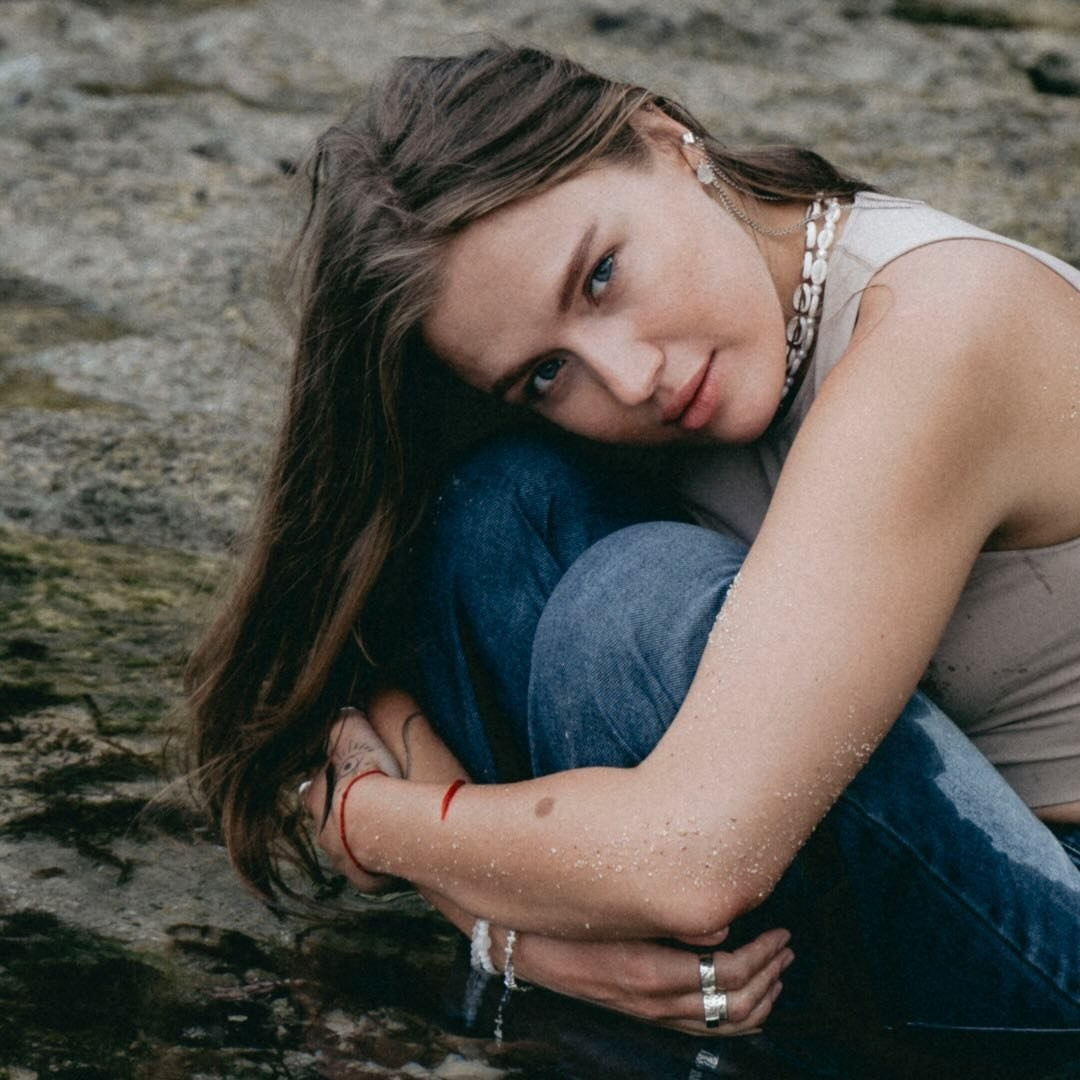 Женщина сидит у воды на каменистой местности в джинсах и майке без рукавов, с задумчивым выражением лица.