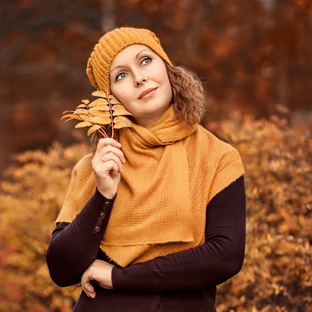Девушка в горчичном шарфе и шапке держит листья рябины в руке и смотрит, задумавшись, вдаль.