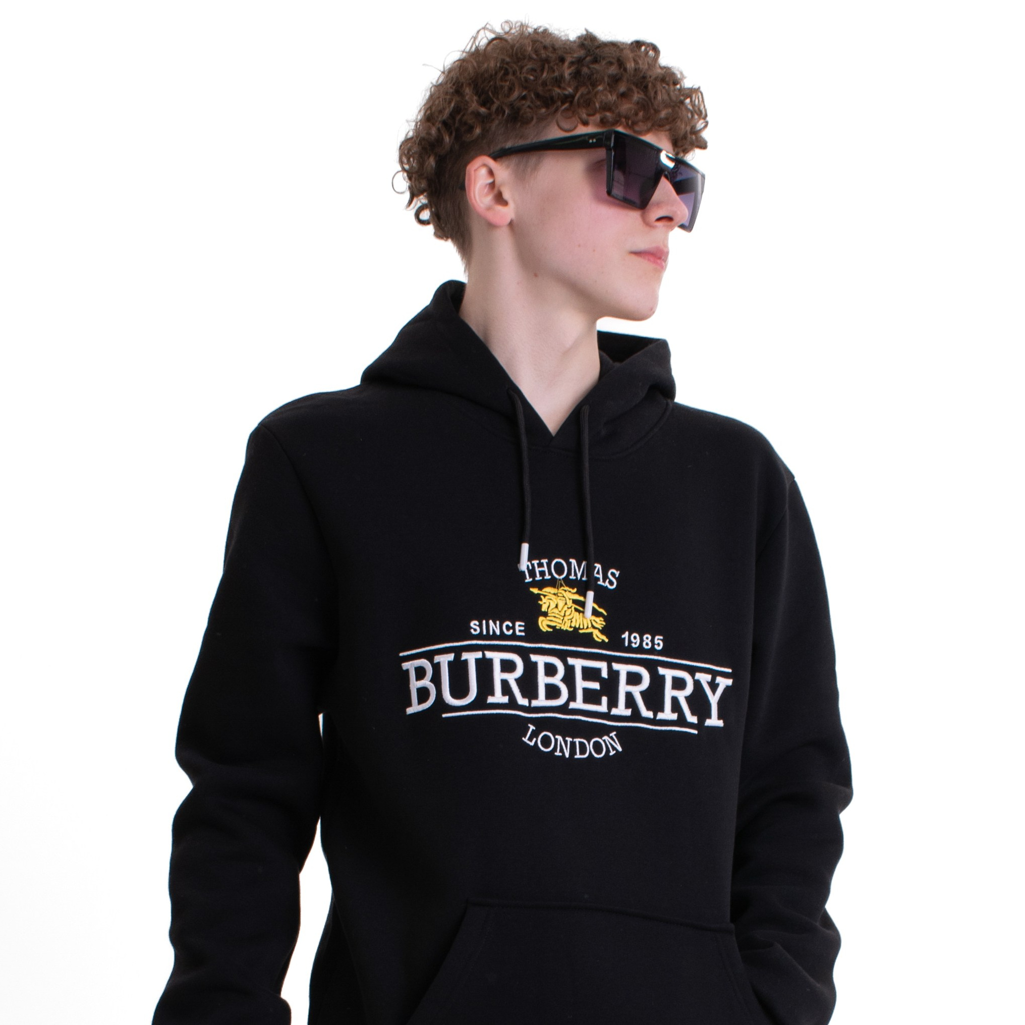 Кудрявый парень стоит на белом фоне в очках и черном худи