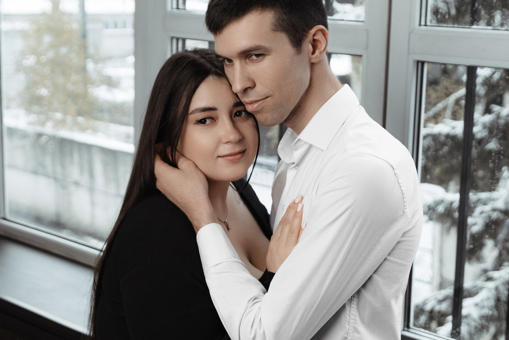 Пара обнимается у окна, мужчина стоит, женщина прижата к нему.