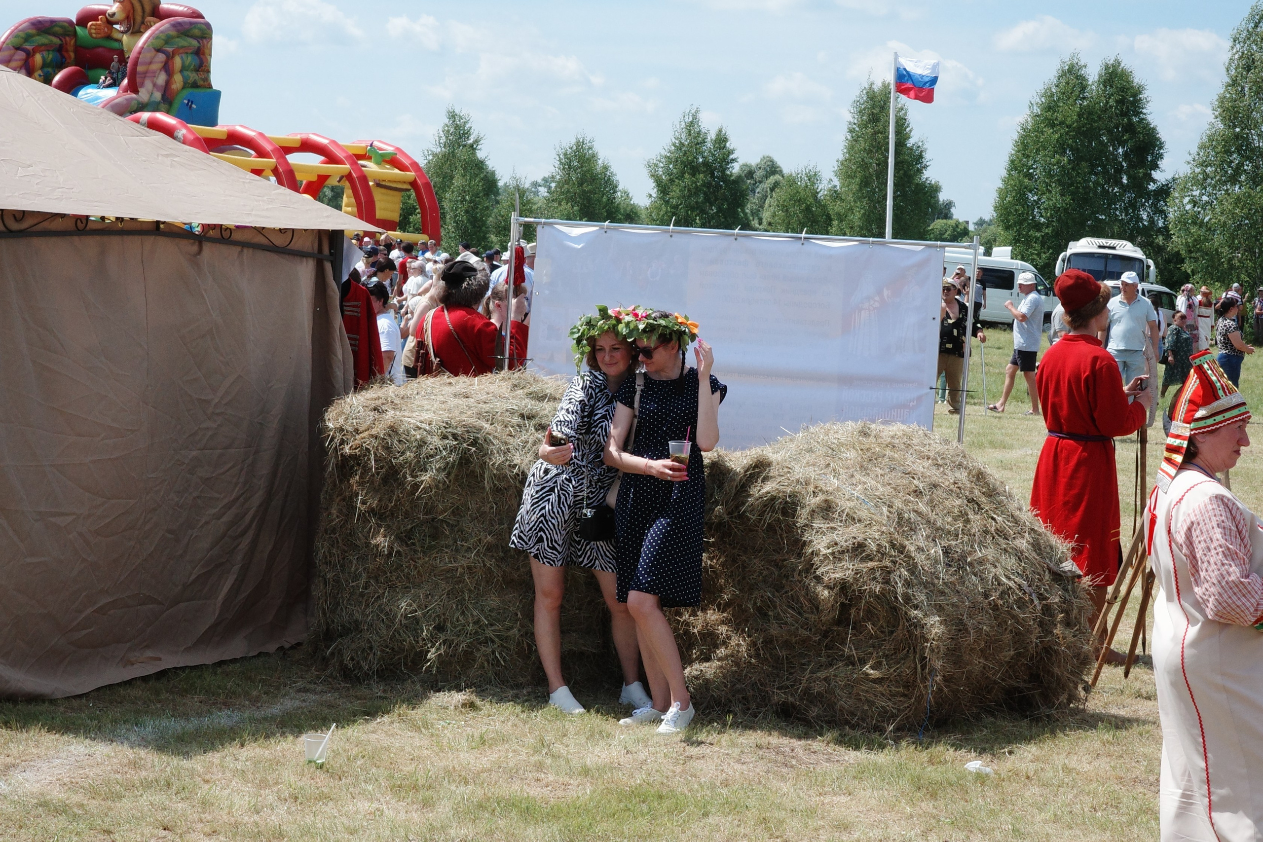 Две девушки в летних платьях с цветочными венками опираются на тюки сена и смеются