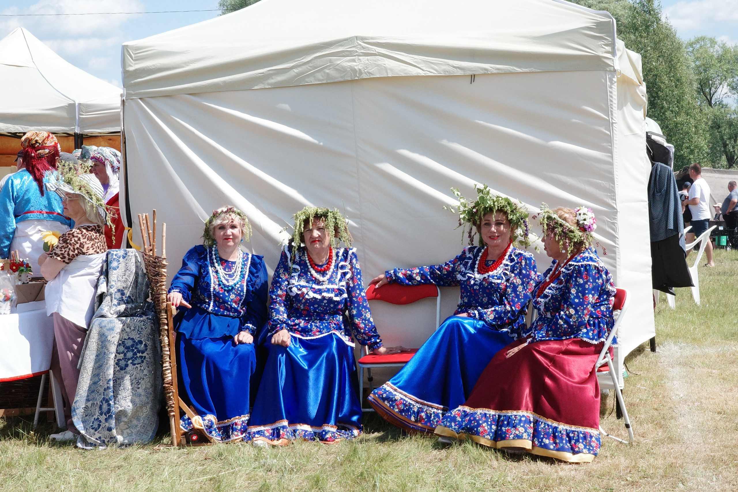 Четыре женщины в синих национальных костюмах с цветочными венками на головах сидят в ряд