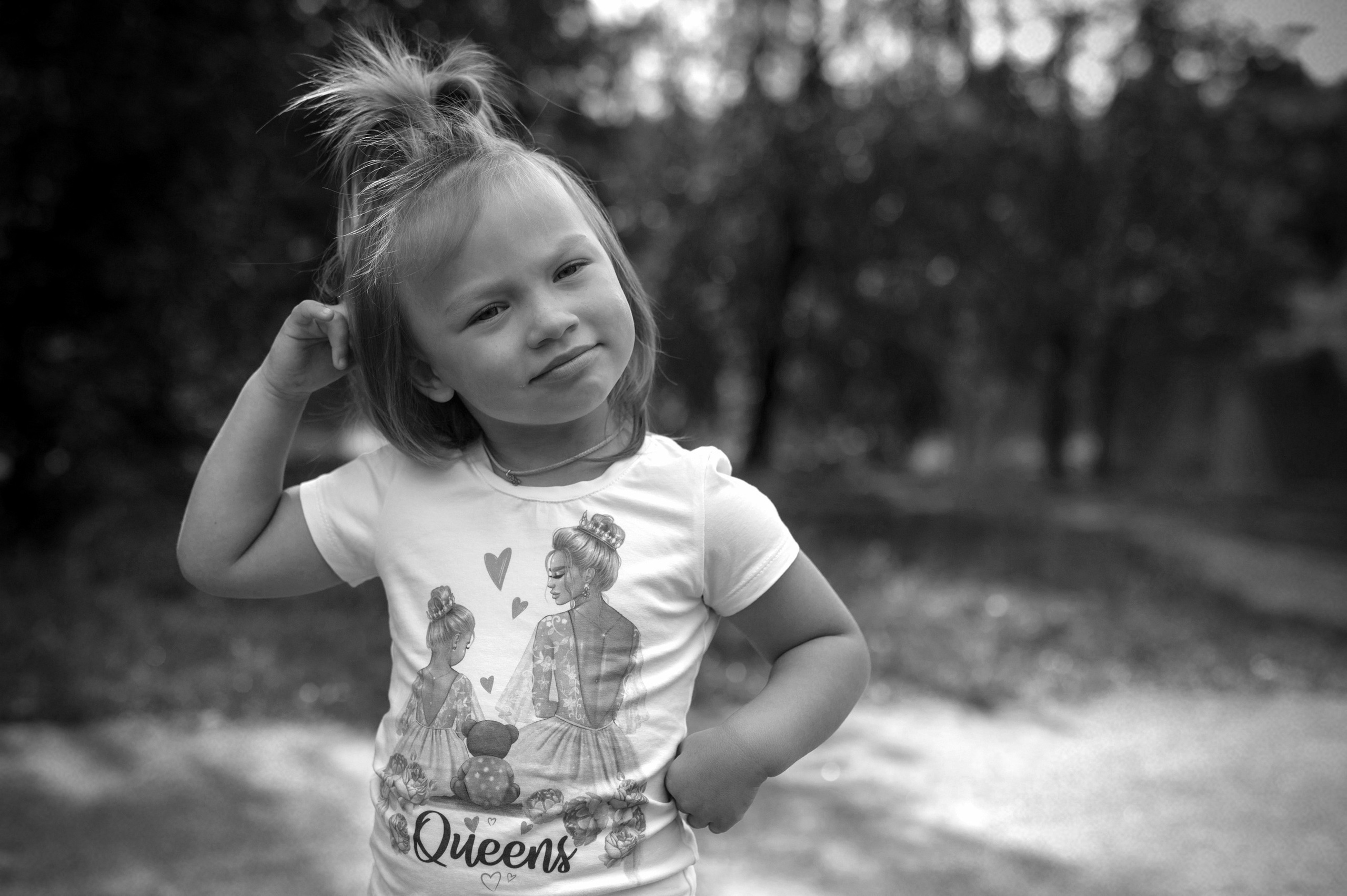 Маленькая девочка с хвостиком на голове в белой футболке с надписью "Queens"