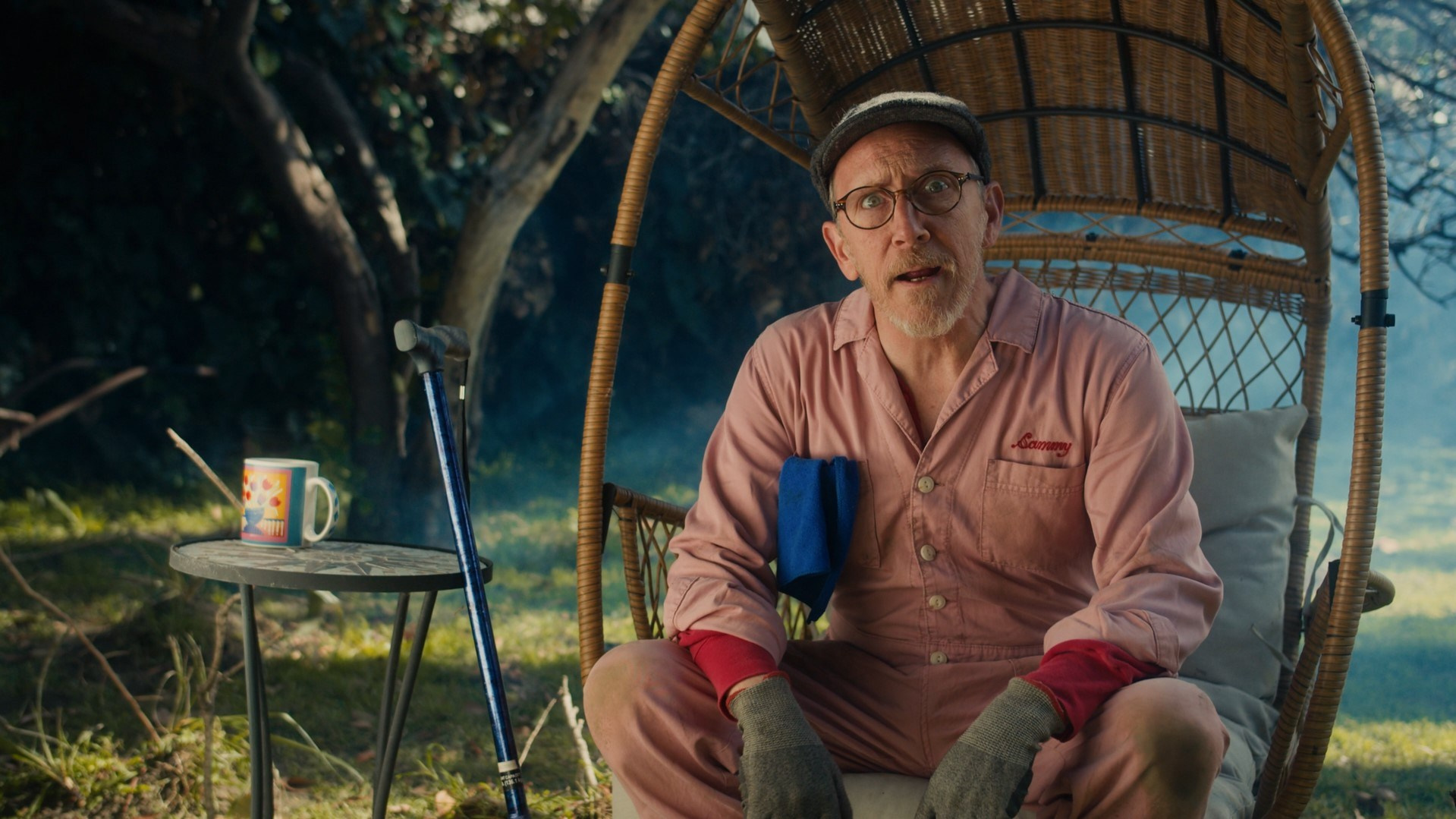 Elderly man sitting in a garden chair, holding a cup and cane, set in a peaceful outdoor scene.