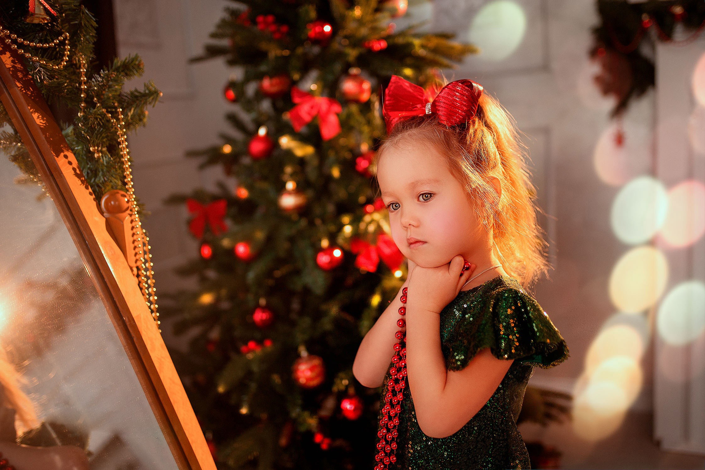Девочка с красным бантом у новогодней ёлки стоит перед зеркалом