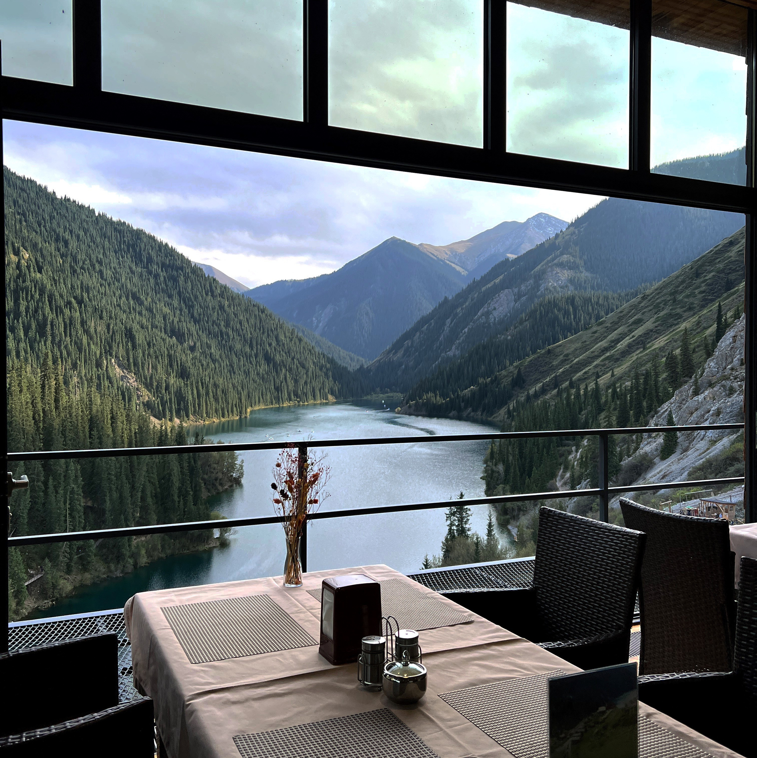 Ярко-бирюзовое озеро Иссык недалеко от Алматы окружено густыми хвойными лесами на фоне высоких гор. На переднем плане видны деревья и травянистая растительность, подчёркивающие природную красоту пейзажа. Чистое голубое небо добавляет яркости сцене.