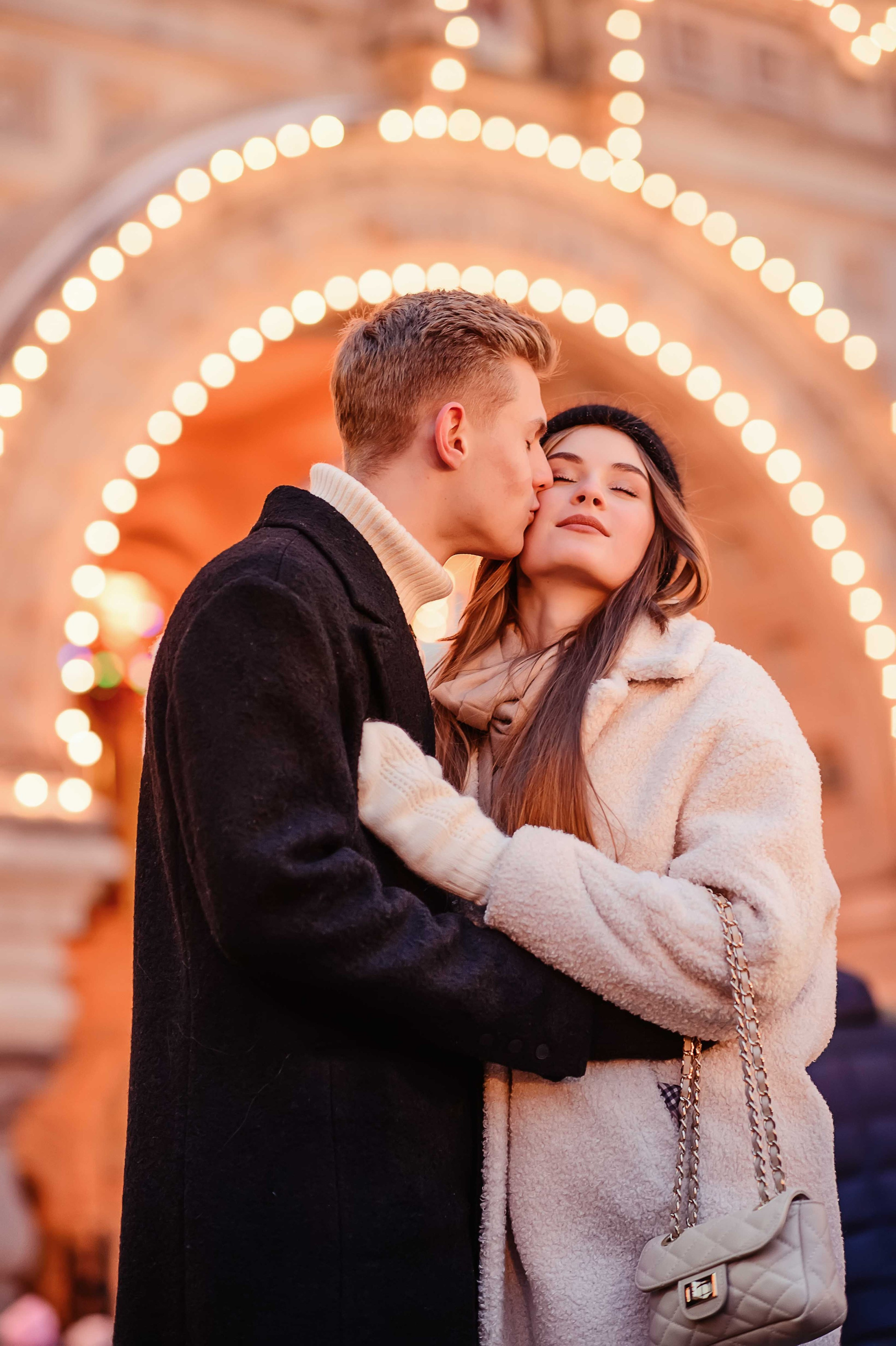 Фотография, на которой изображена пара влюбленных молодых людей;  парень целует девушку в щеку на фоне ГУМа на Красной площади в Москве