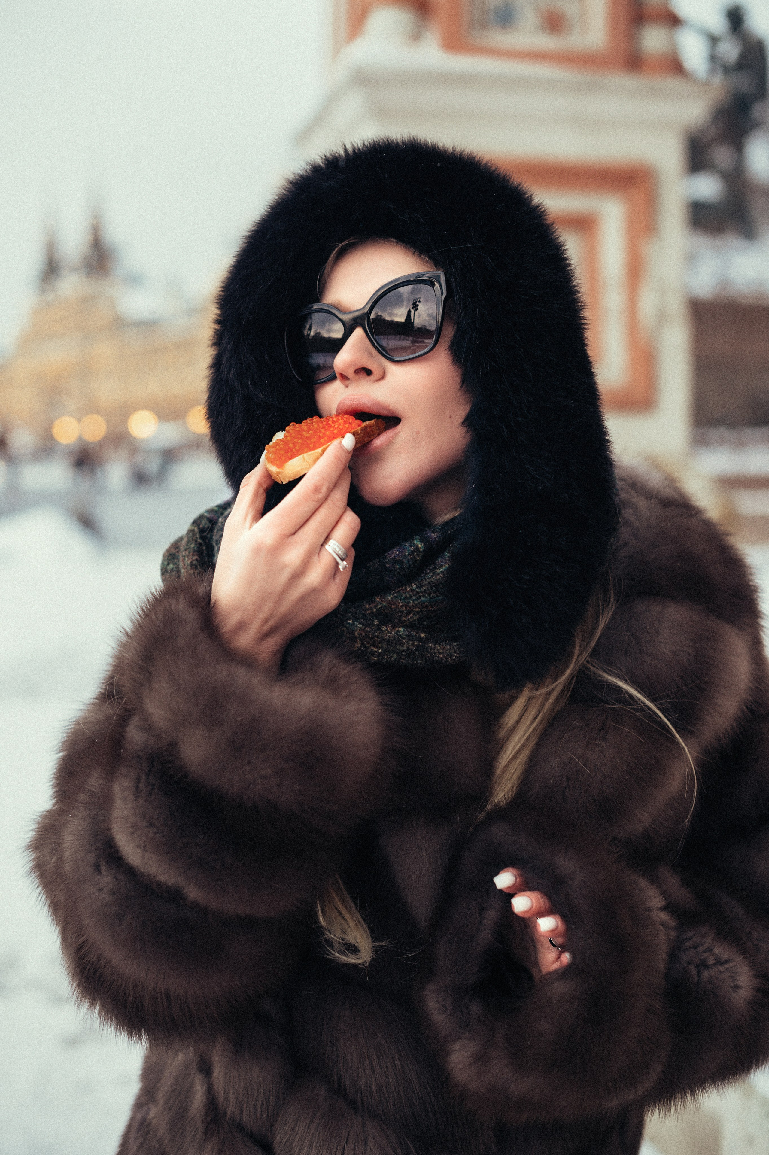 Девушка в соболиной шубе и меховом платке, в темных очках на Красной площади стоит с бутербродом с красной икрой и готовится его съесть