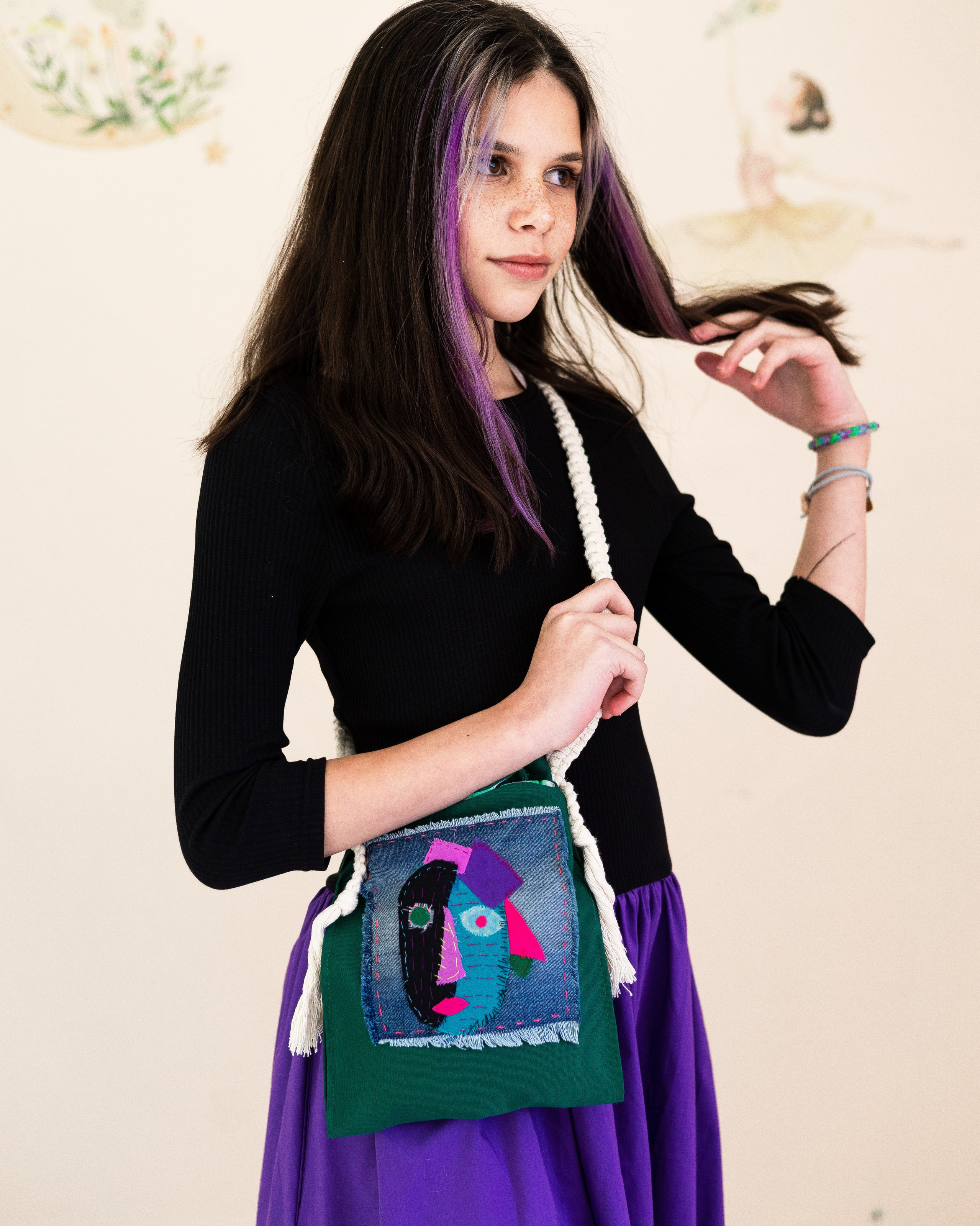 Сумку кросс-боди с авторским принтом, выполненным в технике аппликация, и ручками, связанными в технике макраме, показывает девочка 11 лет