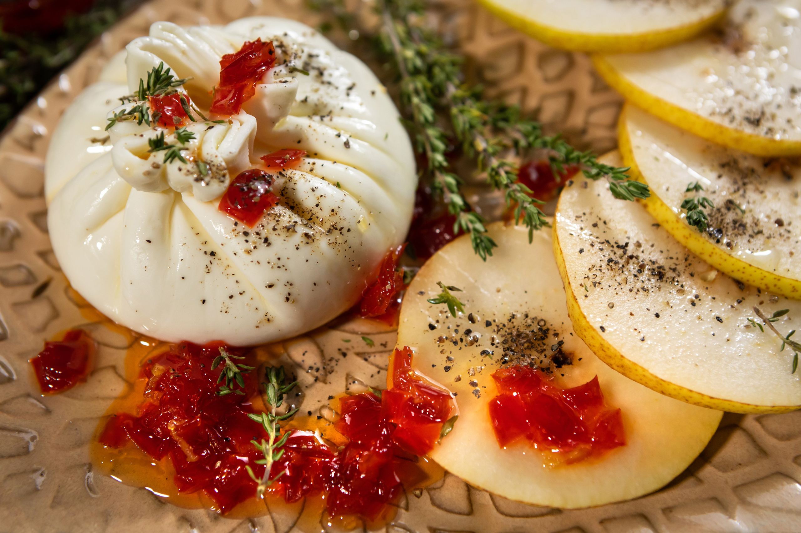 Разрезанный мешочек сыра буррата на белой прямоугольной тарелке с помидорками черри, зеленью и специями, рядом листья красного салата.
