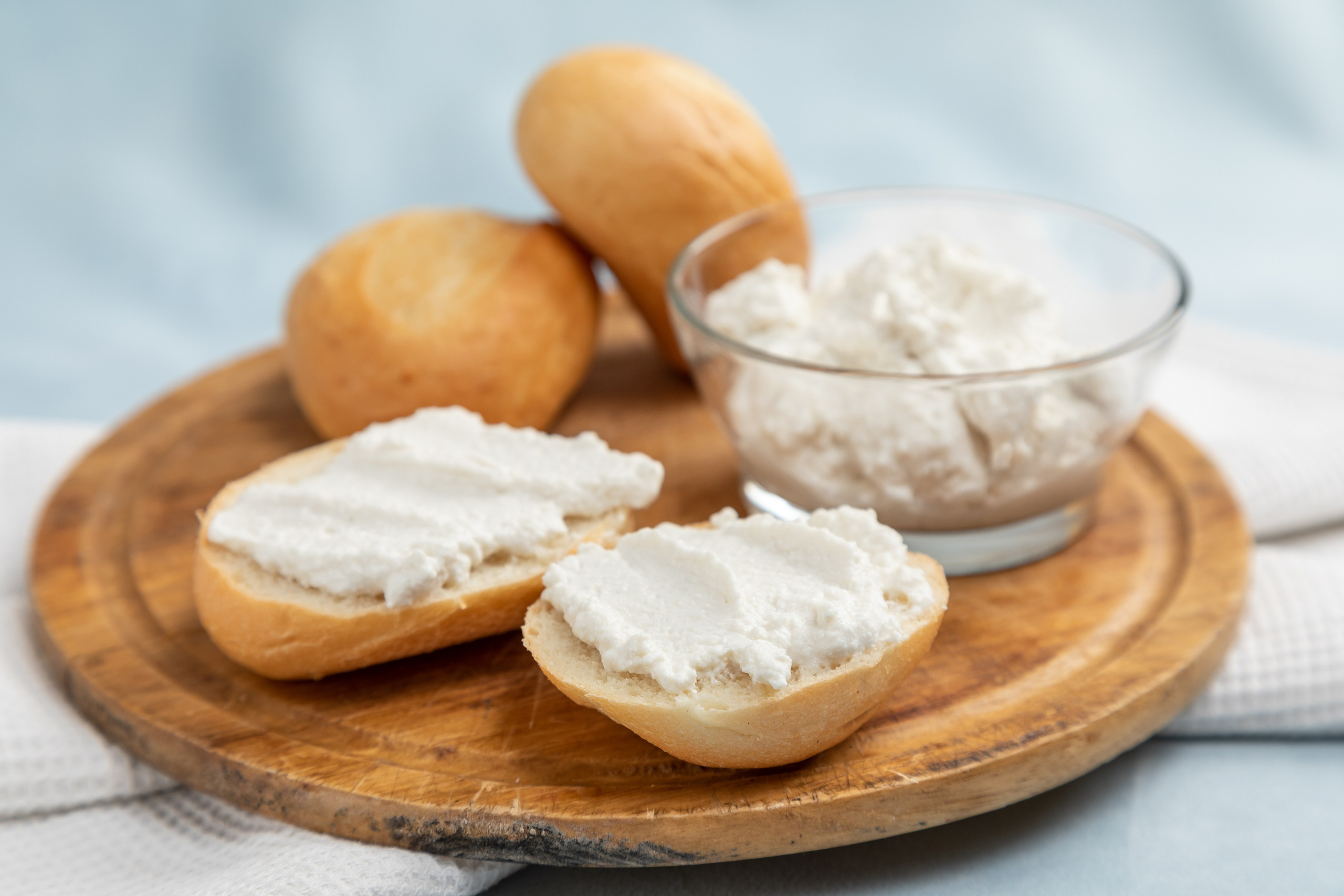 Два бутерброда с сыром рикотта и стеклянная мисочка с тем же сыром на круглой досочке, на голубой скатерти.