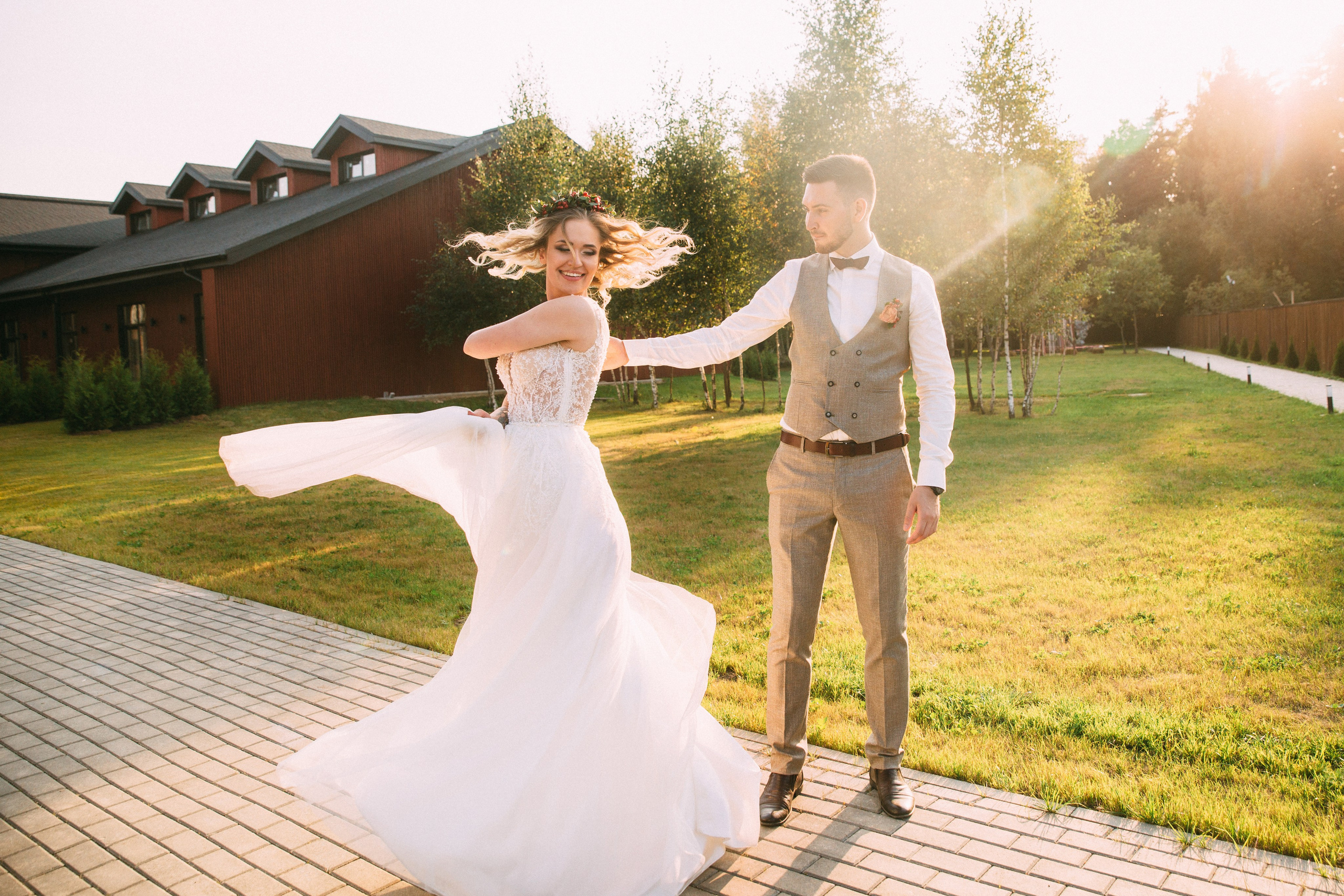Невеста в свадебном платье кружится на фоне коттеджного поселка в Подмосковье, жених нежно держит её за руку. Романтичная свадебная фотография на природе.