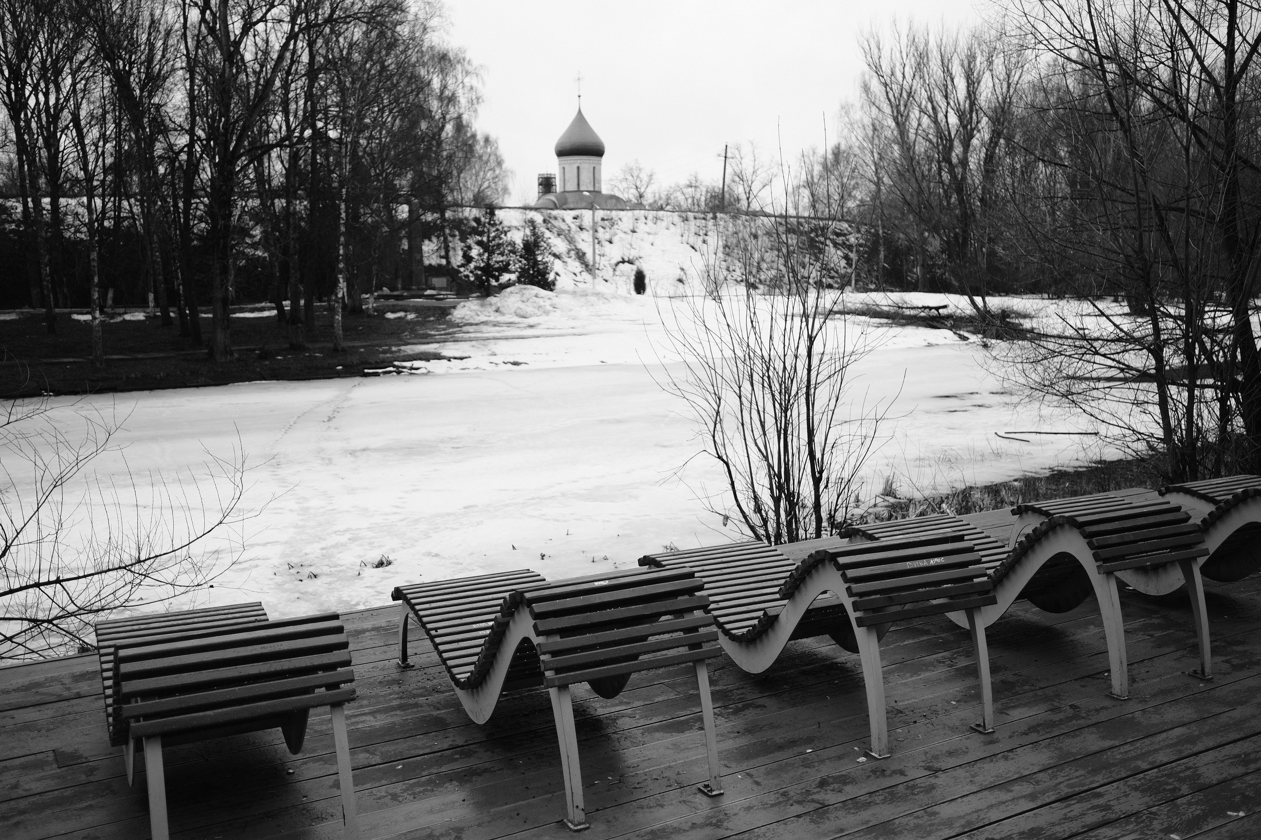 Волнообразные скамейки на набережной у замерзшей реки с церковью на горизонте, голые деревья, черно-белое зимнее фото