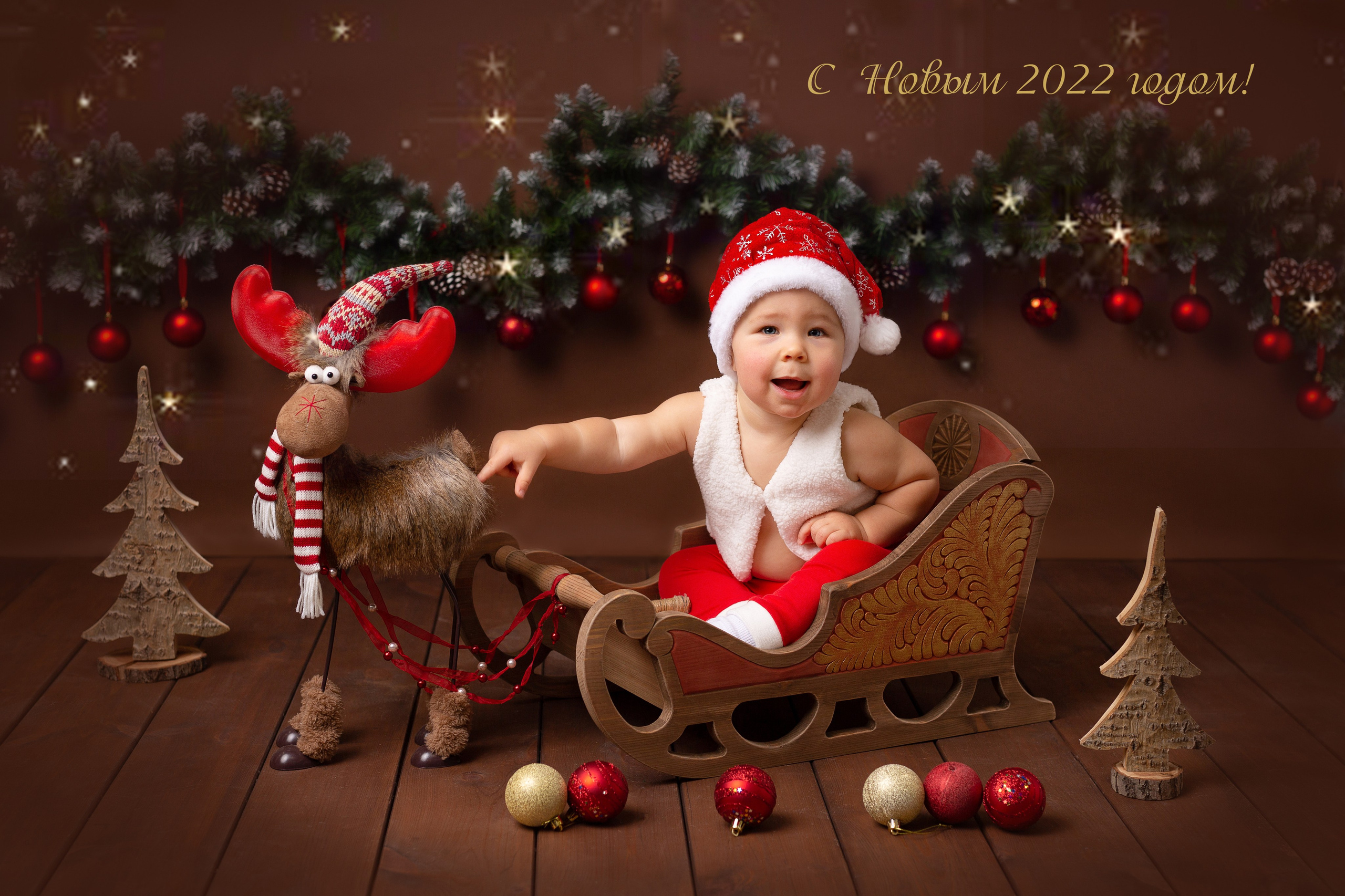 Ребёнок в красных штанах и колпаке деда мороза сидит в новогодних санях.