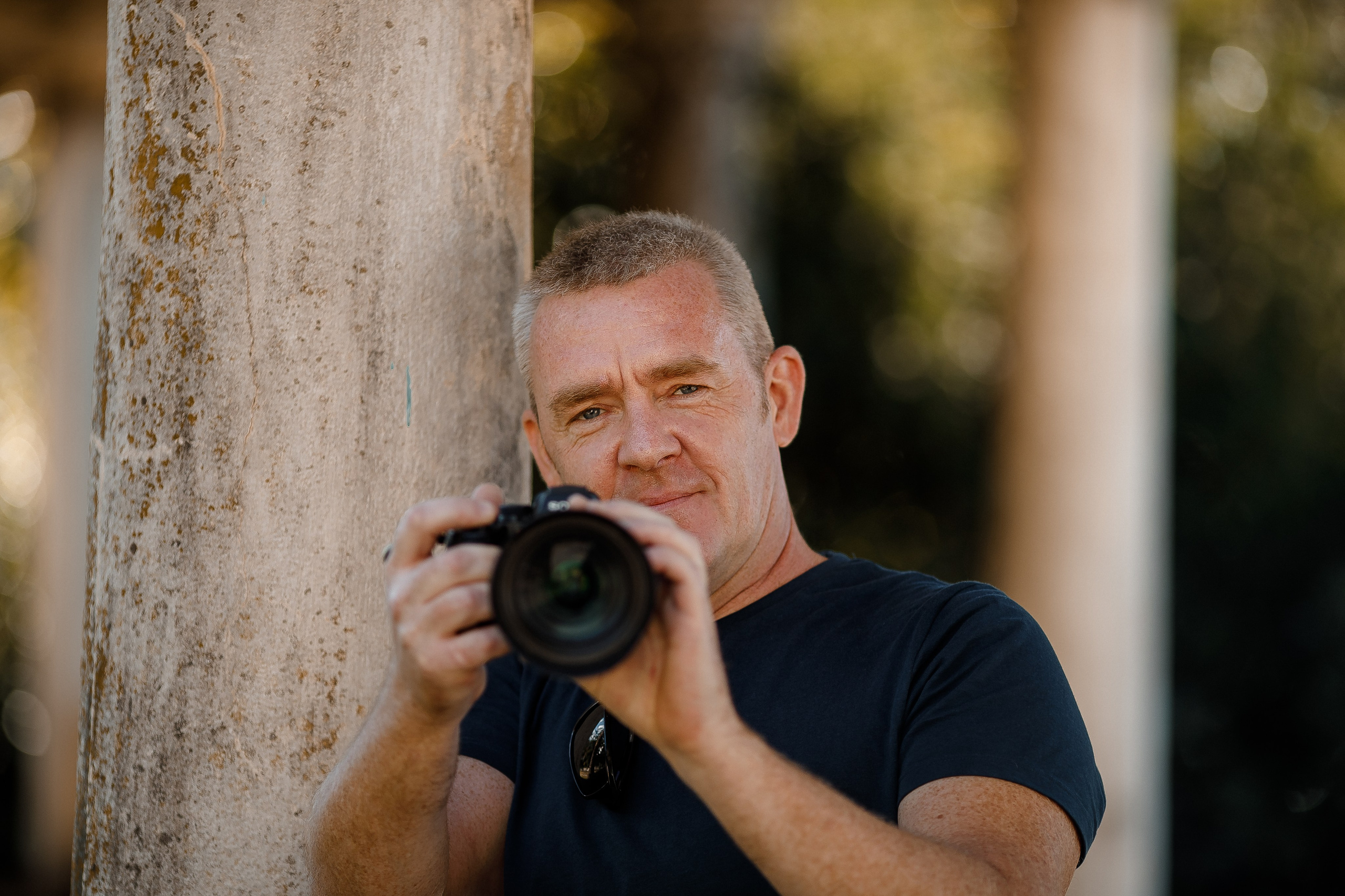 Мужчина с короткими светлыми волосами держит фотоаппарат и смотрит прямо в объектив. Он стоит возле колонны на фоне зелени и солнечного света