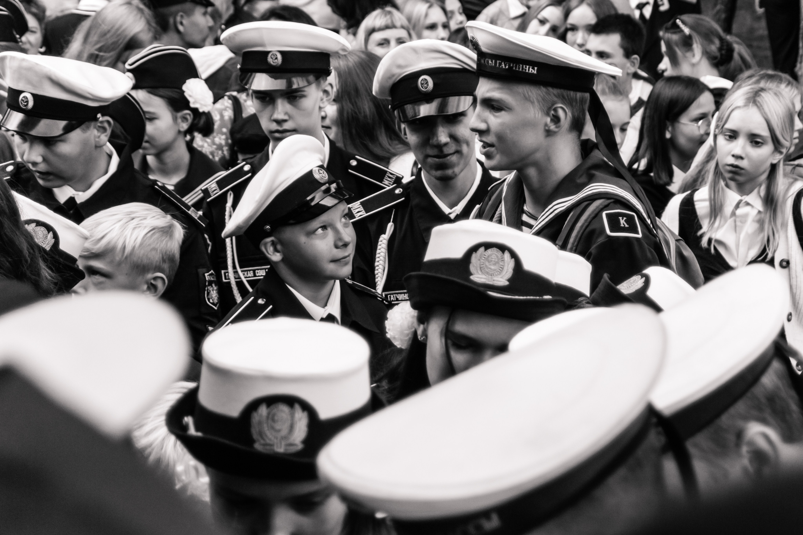 Чёрно-белая фотография группы молодых людей в морской форме с фуражками, стоящих на митинге или празднике. Они сосредоточены на происходящем вокруг. Среди них выделяются несколько подростков, один из которых смотрит вверх с любопытством. Сцена передаёт атмосферу торжественности и ожидания. Парад, кадеты, школьная линейка, 1 сентября, диалог, разговор, встреча, репортаж.