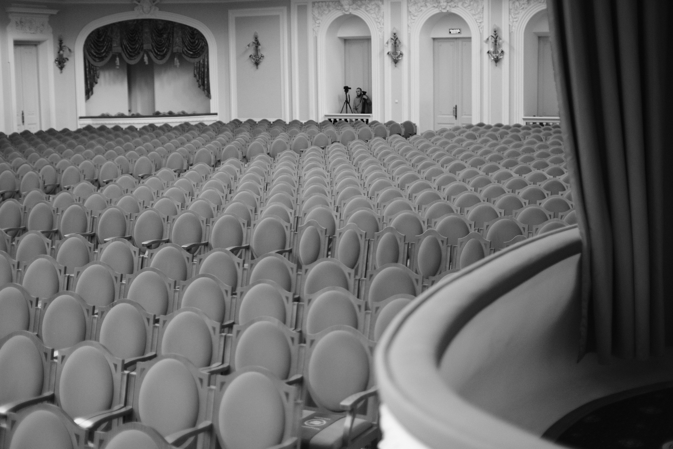 Черно-белая фотография: пустой концертный зал с рядами кресел, орнаментальная архитектура, фотограф на балконе с камерой, интерьер театра