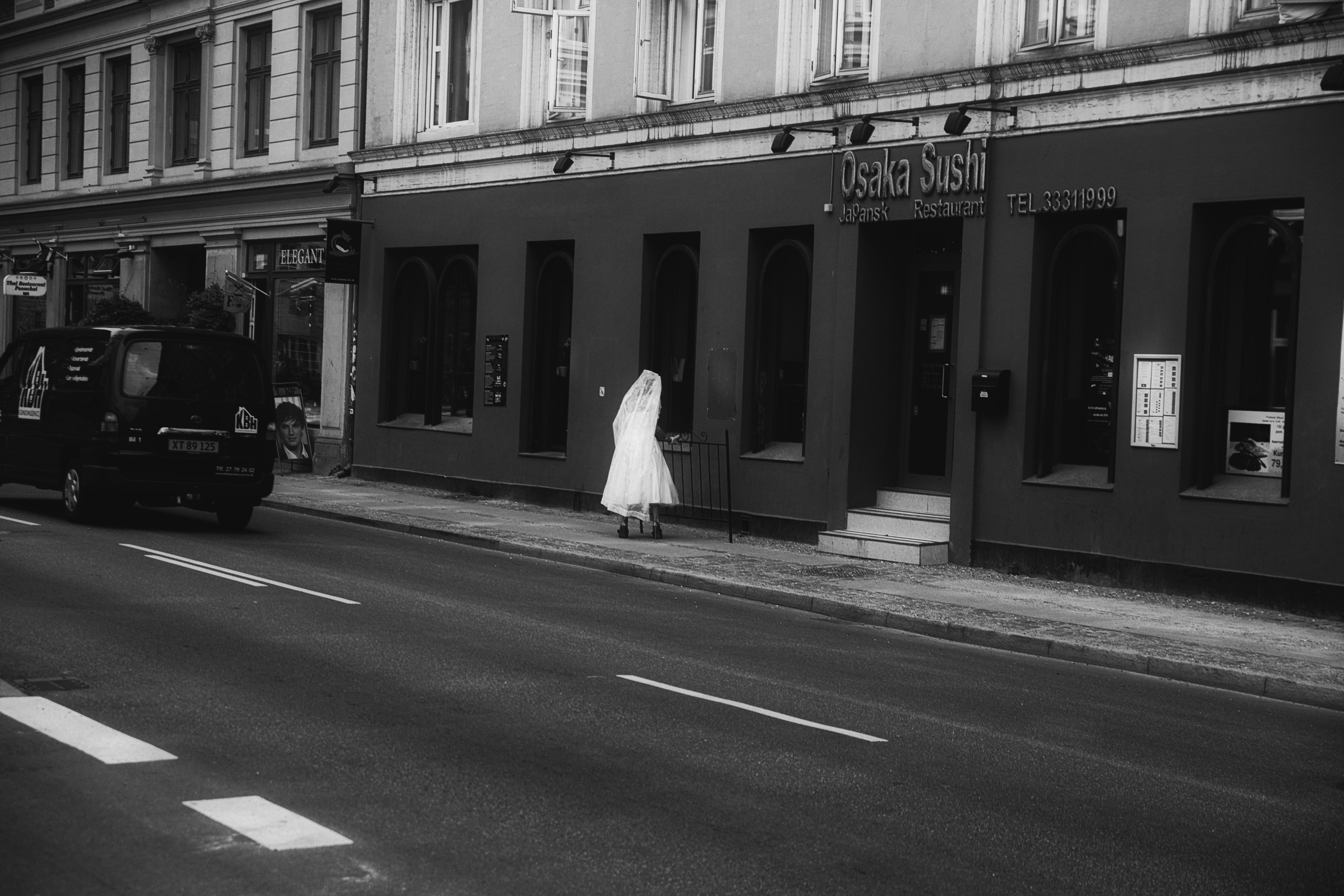 Черно-белая уличная фотография: девушка в белом платье и фате идёт по тротуару мимо ресторана, городской пейзаж, чёрный фургон, документальная съёмка