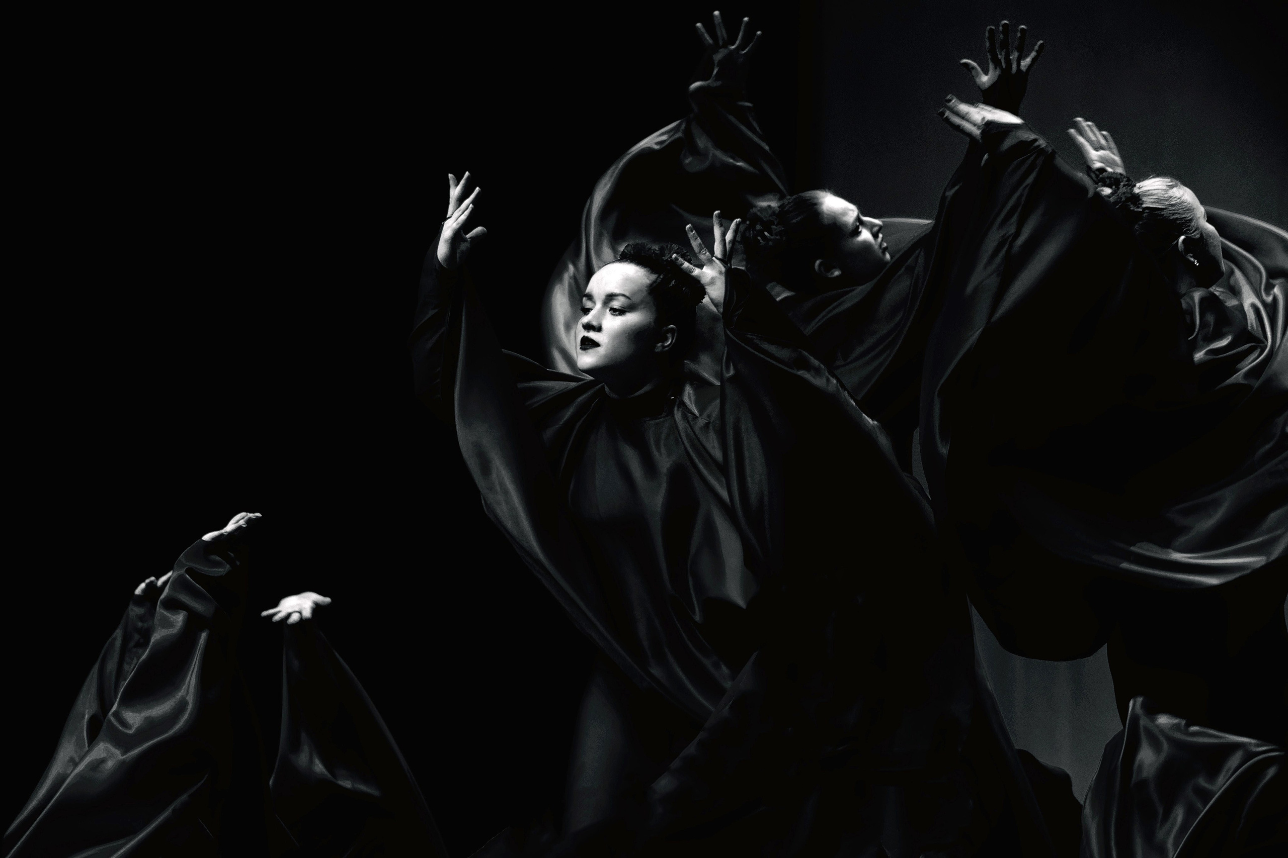 Группа танцоров в черных развевающихся одеждах с поднятыми руками, драматичная хореография, театральное освещение