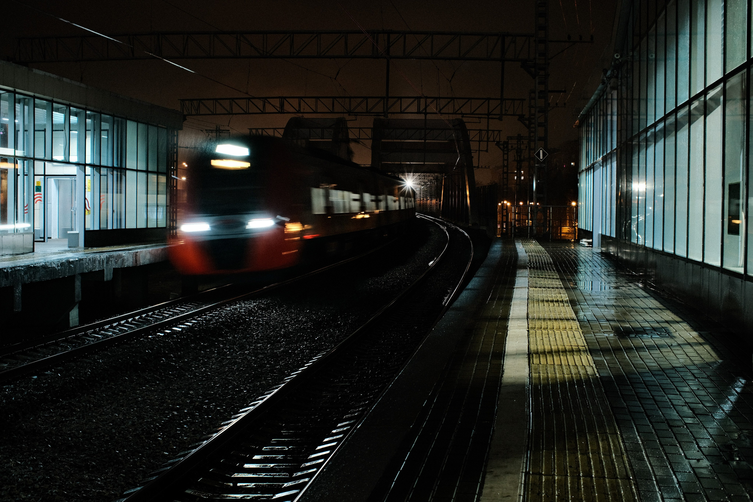 Ночная фотография железнодорожной станции: размытый от движения поезд проезжает мимо платформы, мокрые рельсы и плитка отражают свет, стеклянные павильоны по бокам, городская атмосфера