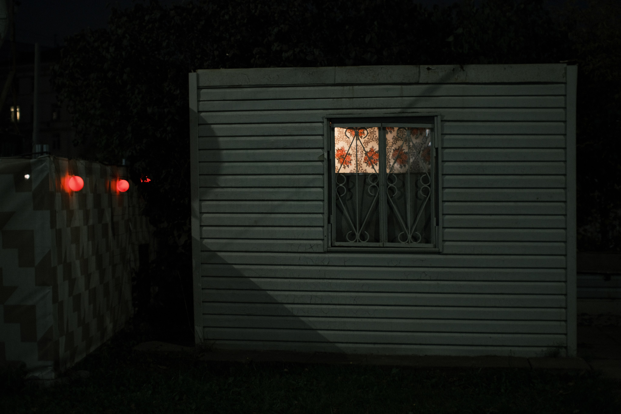 Ночная городская фотография: освещённое окно с узорными шторами и решёткой в деревянном строении, красные огни на заборе слева, тёмная атмосфера