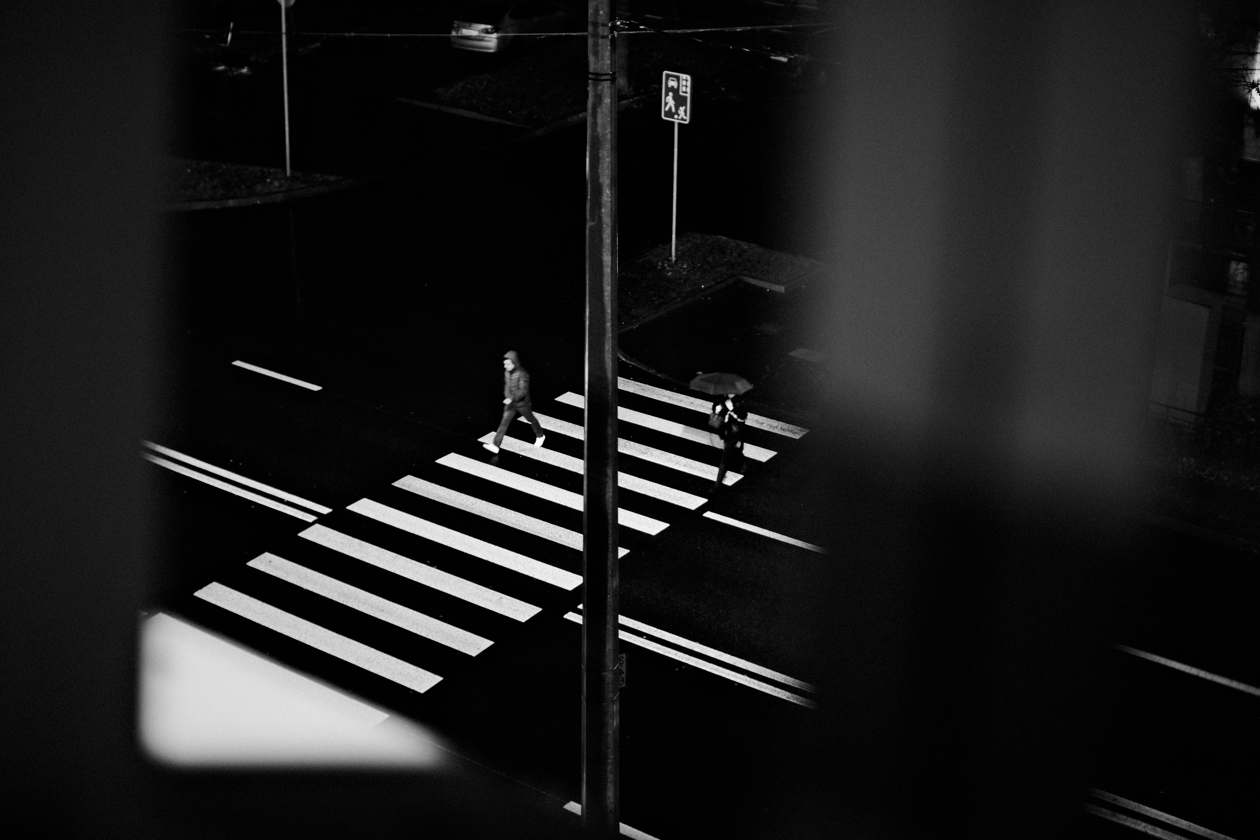 Черно-белая уличная фотография: вид сверху на пешеходный переход, два человека переходят дорогу, один с зонтом, геометрическая композиция, ночной город