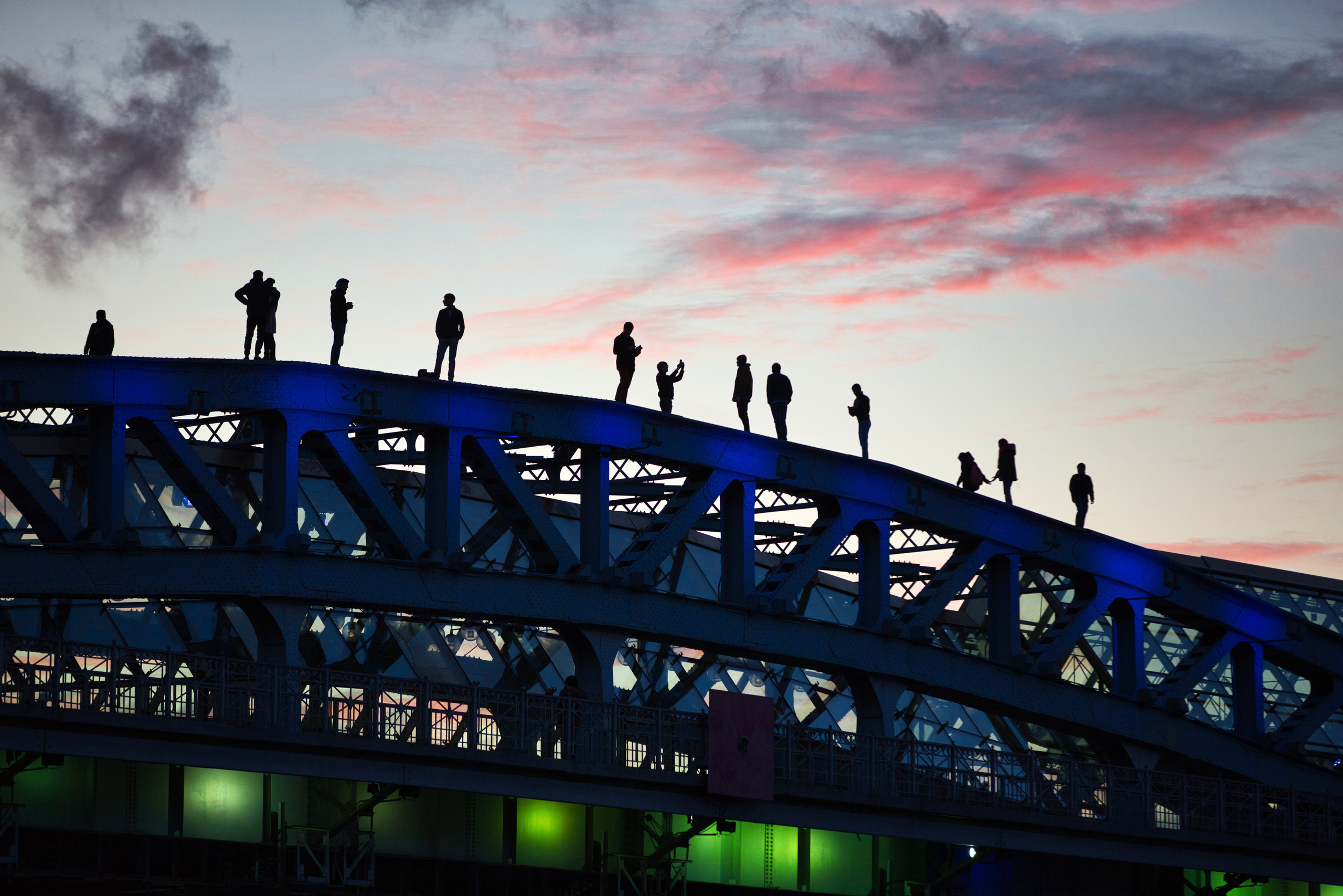 Силуэты людей на мосту на фоне заката: розовые облака, синяя подсветка конструкции, городская фотография, уличный репортаж