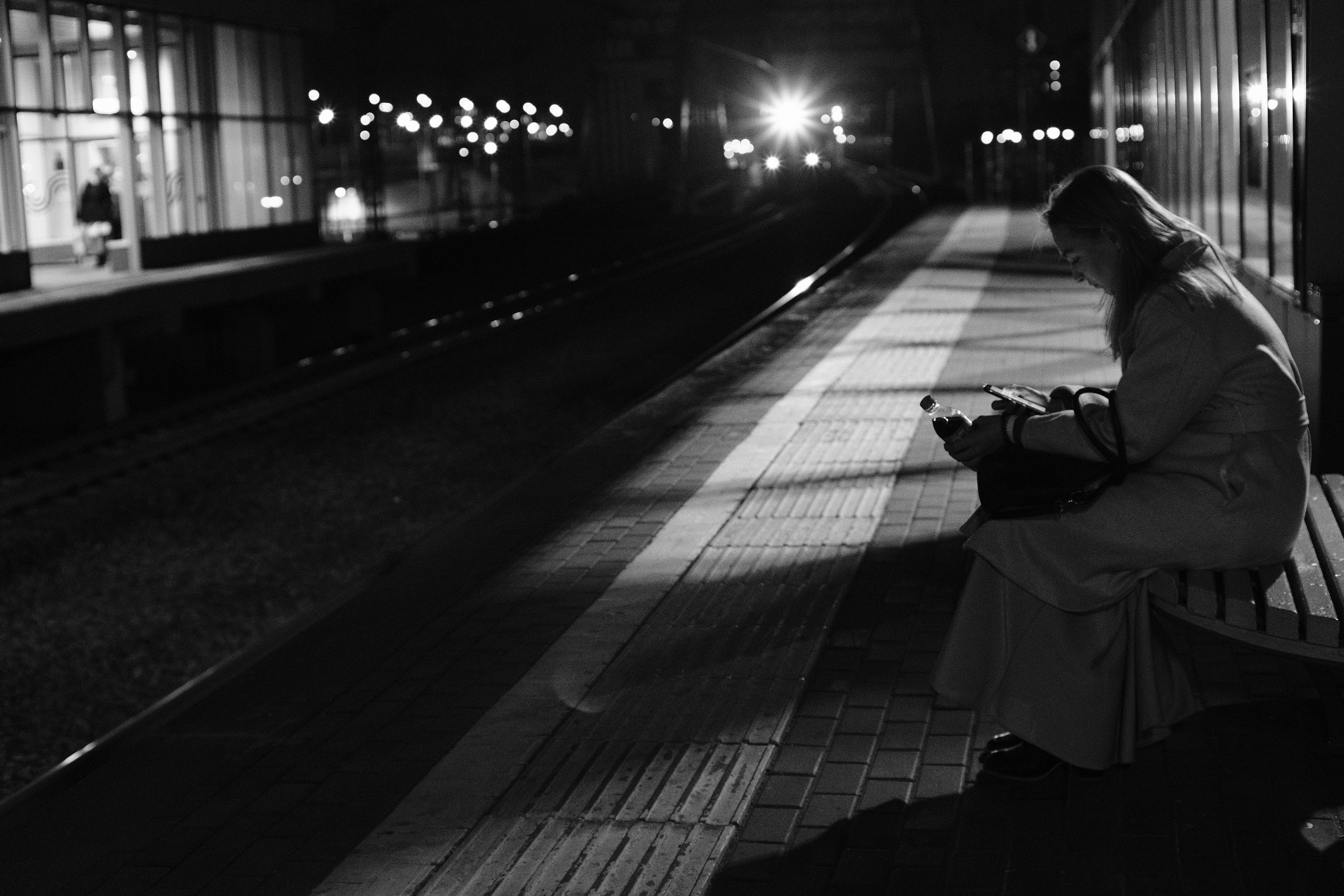 Черно-белая уличная фотография: женщина сидит на скамейке на платформе ночью, смотрит в телефон, огни города на фоне, ожидание транспорта, городской пейзаж