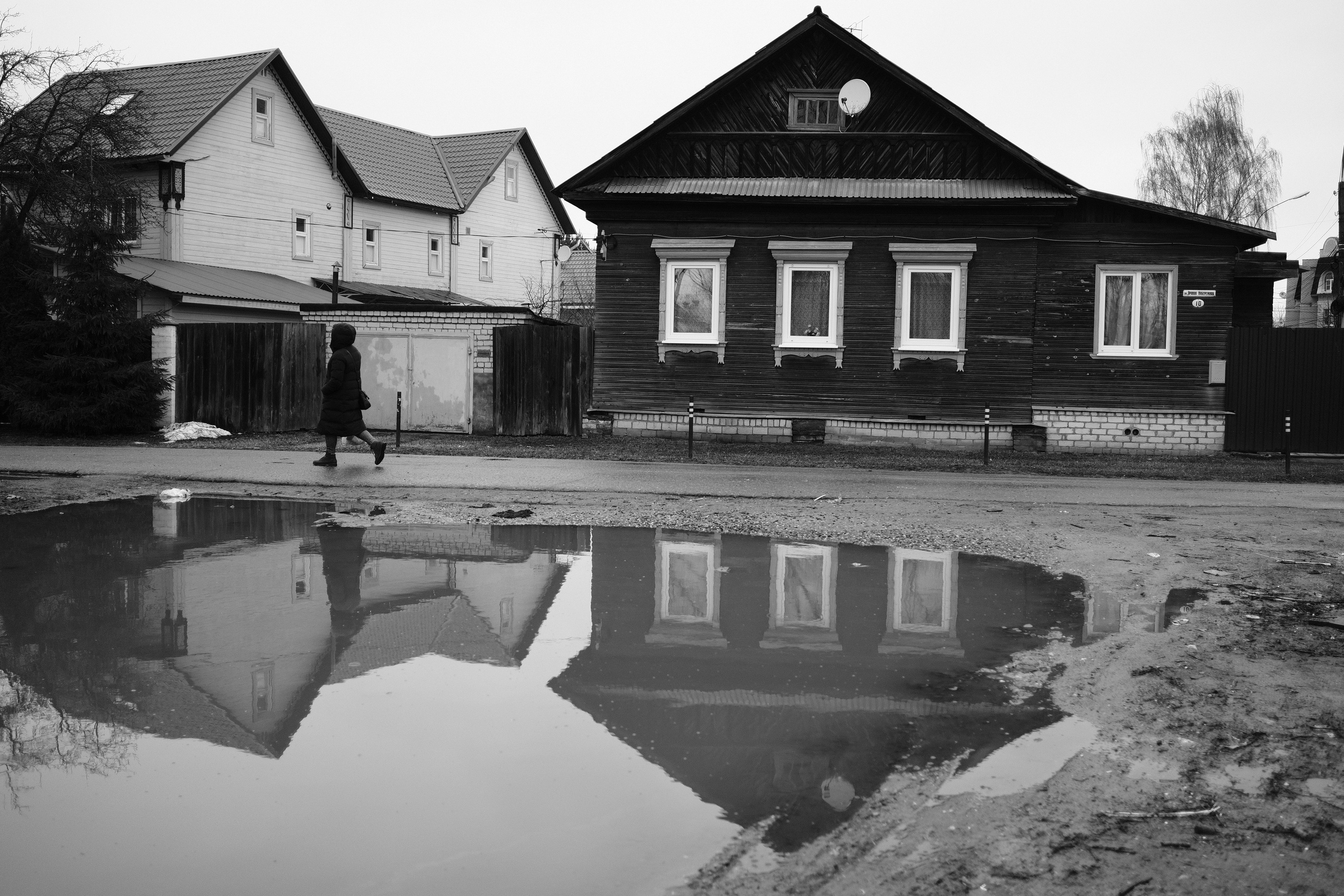 Старый деревянный дом с белыми наличниками отражающийся в луже на улице, человек проходит мимо, контраст старого и нового в Переславле