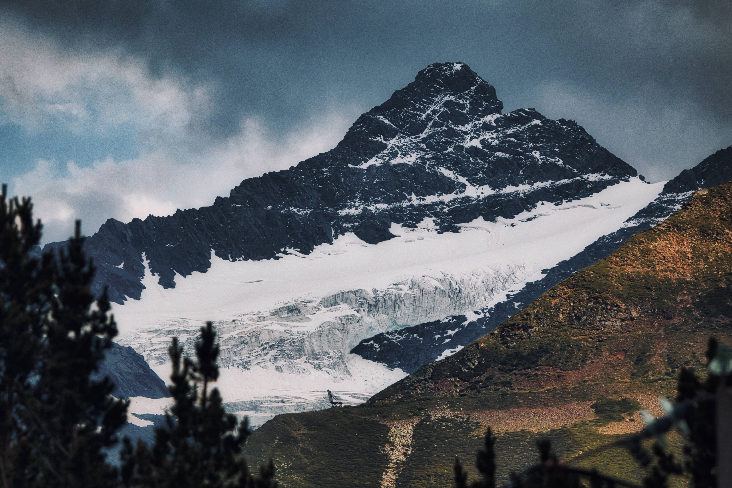 Пейзажная фотография: высокая горная вершина со снегом и ледником, драматичные облака, солнечный свет на склоне, силуэты деревьев на переднем плане