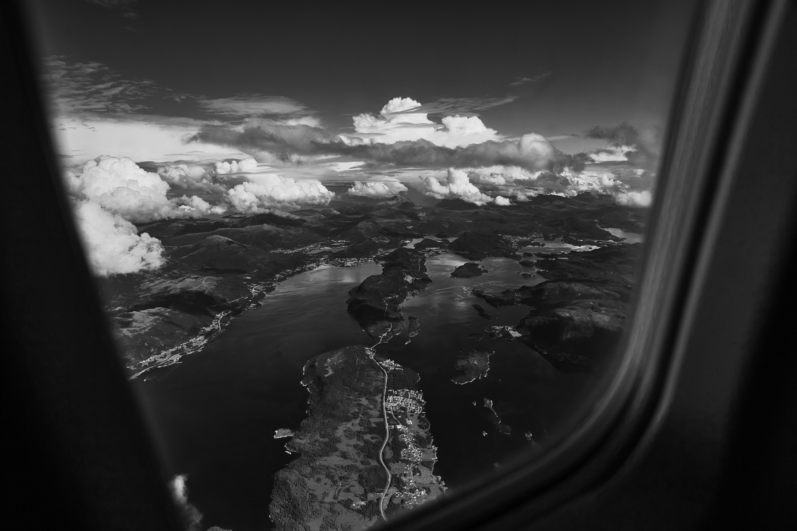 Черно-белая аэрофотосъёмка: вид из иллюминатора самолёта на фьорды и острова, облака, горы, прибрежный пейзаж, Норвегия