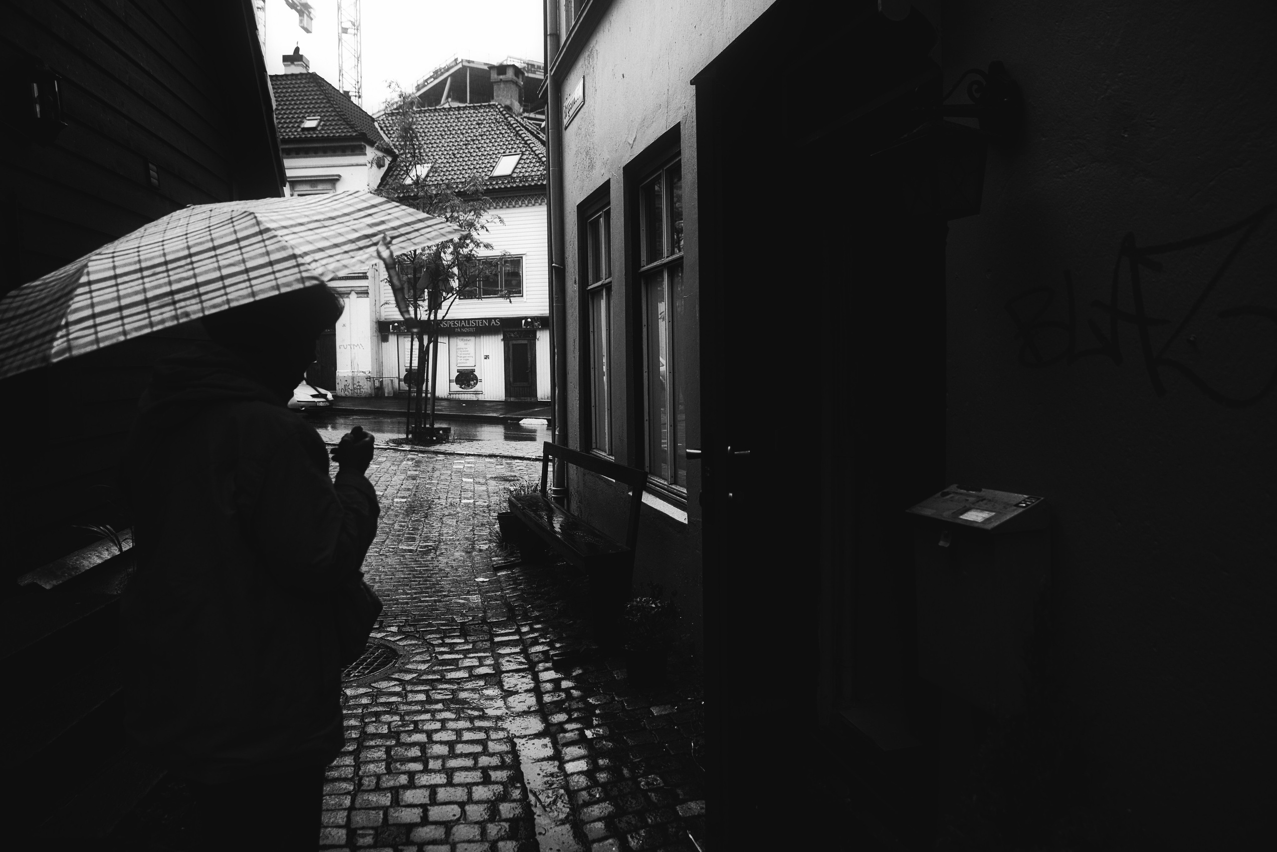 Черно-белая уличная фотография: человек с зонтом в дождливом переулке, мокрая брусчатка, старинные здания, атмосферное фото, городской пейзаж