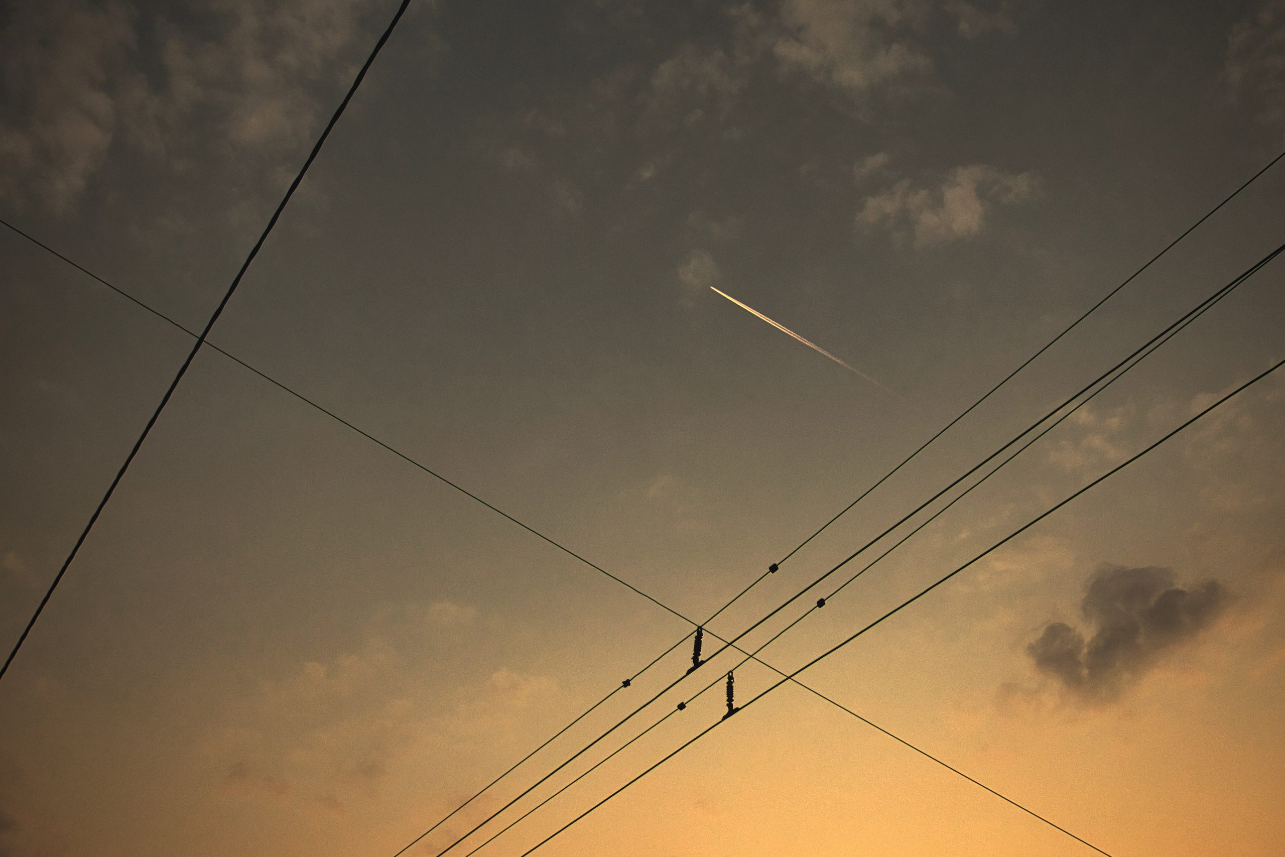 Минималистичная фотография заката: диагональные линии электропередач пересекают небо с теплым оранжевым оттенком, белый конденсационный след самолета на фоне облаков, геометрическая композиция