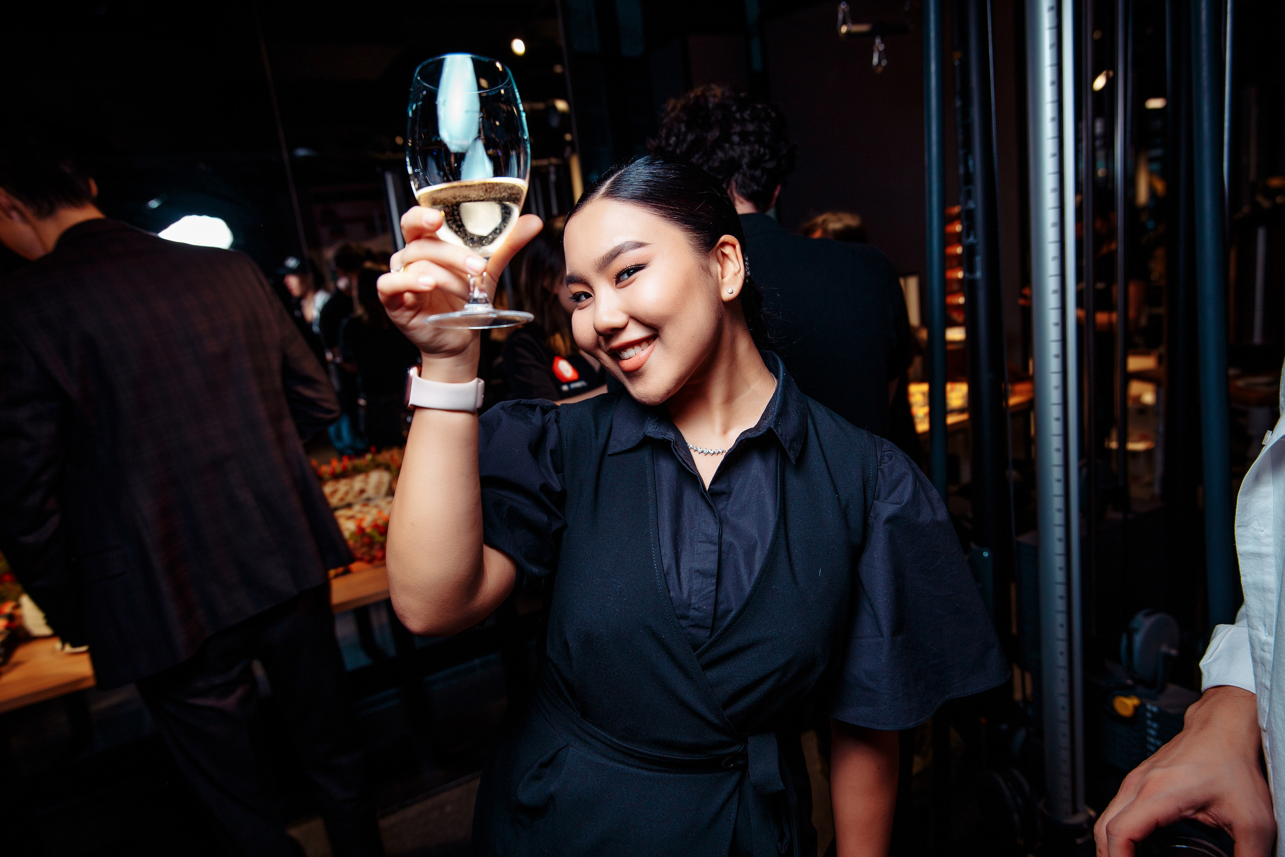 Фотография снятая на мероприятие. Девушка с шампанским в руке, она поднимает его высоко над головой и улыбается.