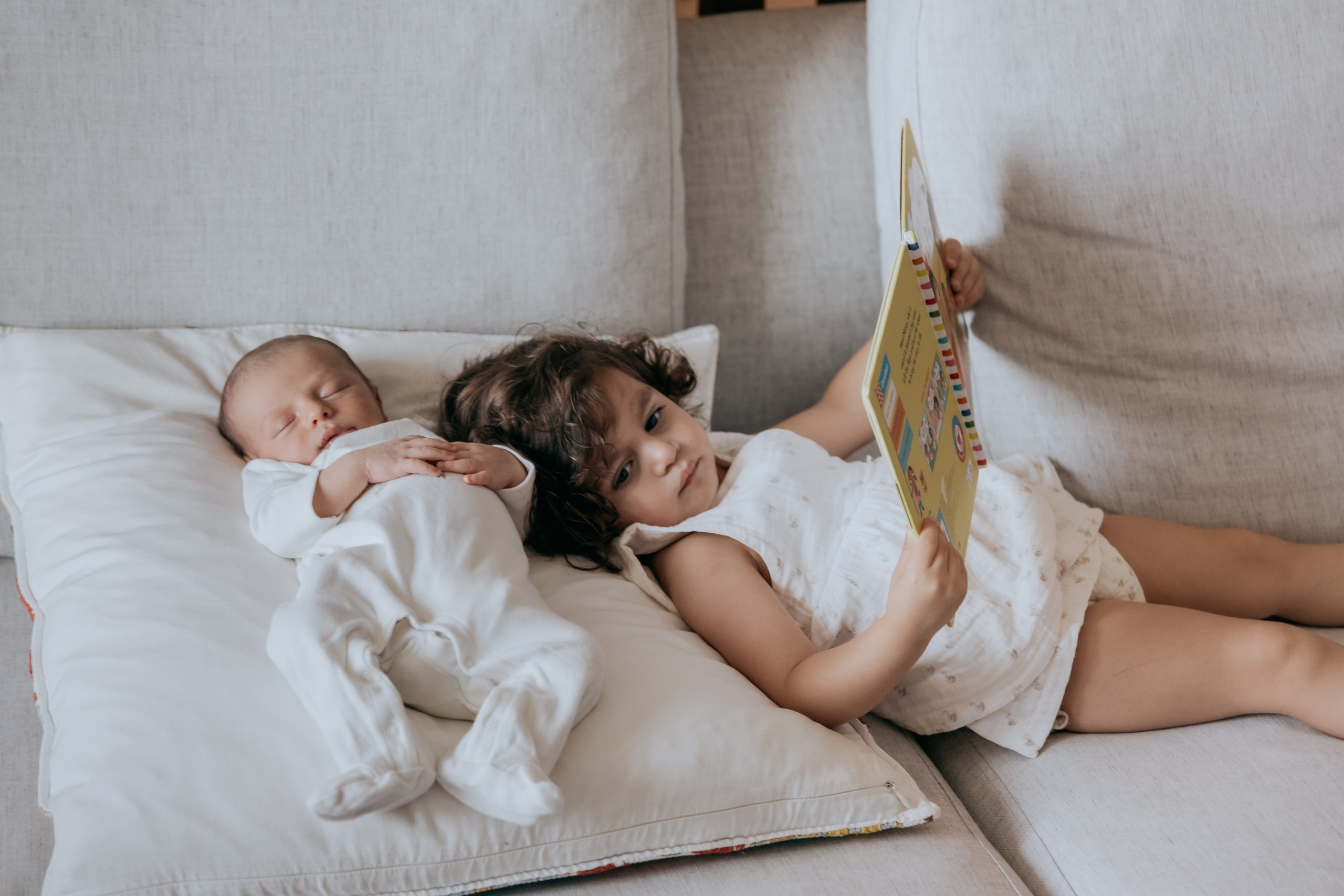 Young girl reading a book next to her sleeping newborn sibling on a couch