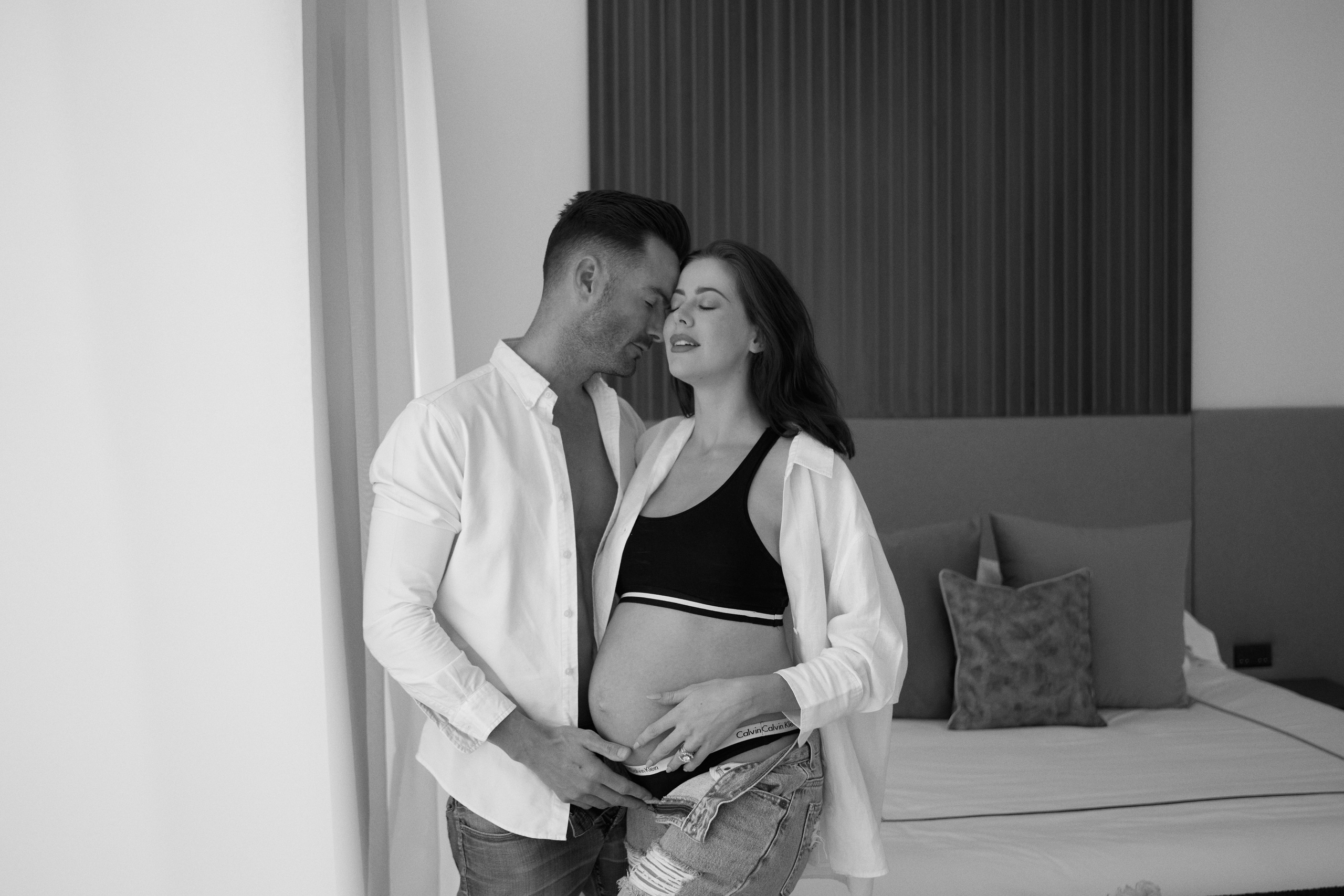 A couple shares an intimate moment, with the man gently holding the woman's pregnant belly in a serene bedroom setting