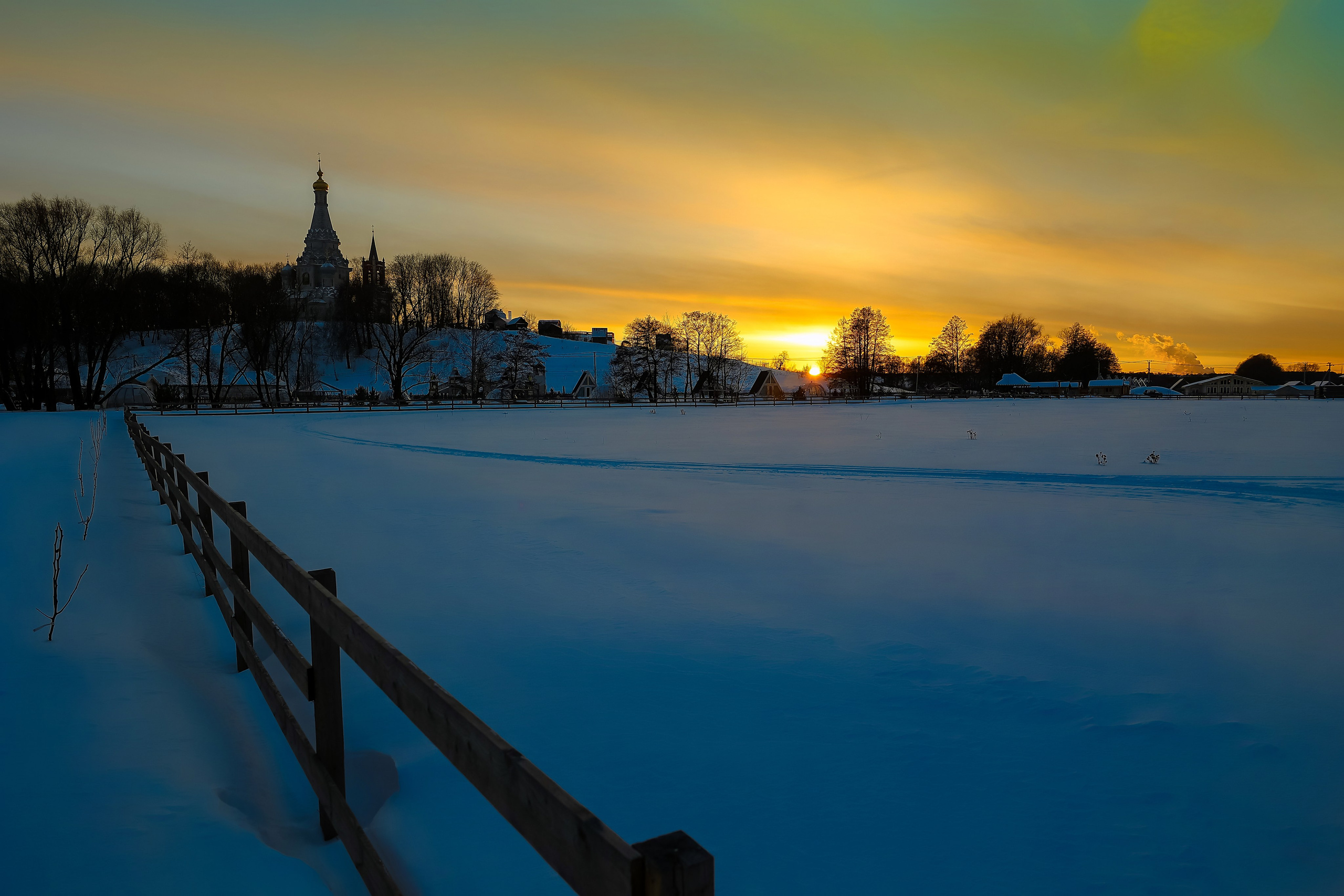 Вечерний зимний пейзаж: заснеженная дорога уходит вдаль к силуэту храма, небо с жёлтыми и бирюзовыми оттенками постепенно темнеет.