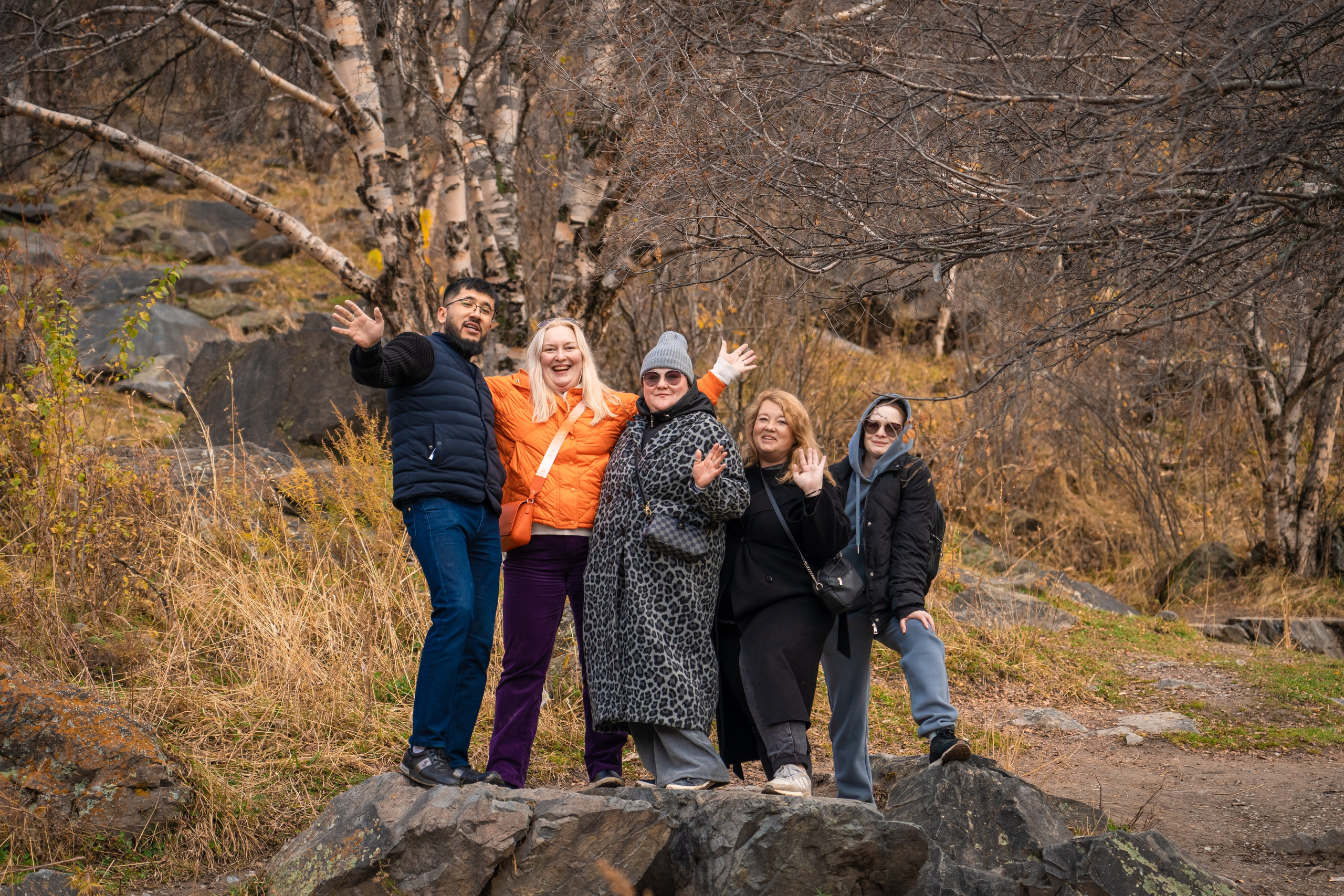 Группа из четырёх человек улыбается и позирует на фоне деревянной арки с надписью "Медвежий водопад". Люди выглядят счастливыми, одеты в светлую и спортивную одежду, очевидно, находятся в походе. За аркой видны склоны с зеленью и каменистая тропа. Атмосфера весёлая и дружелюбная, подчеркивает любовь к природе и приключениям.