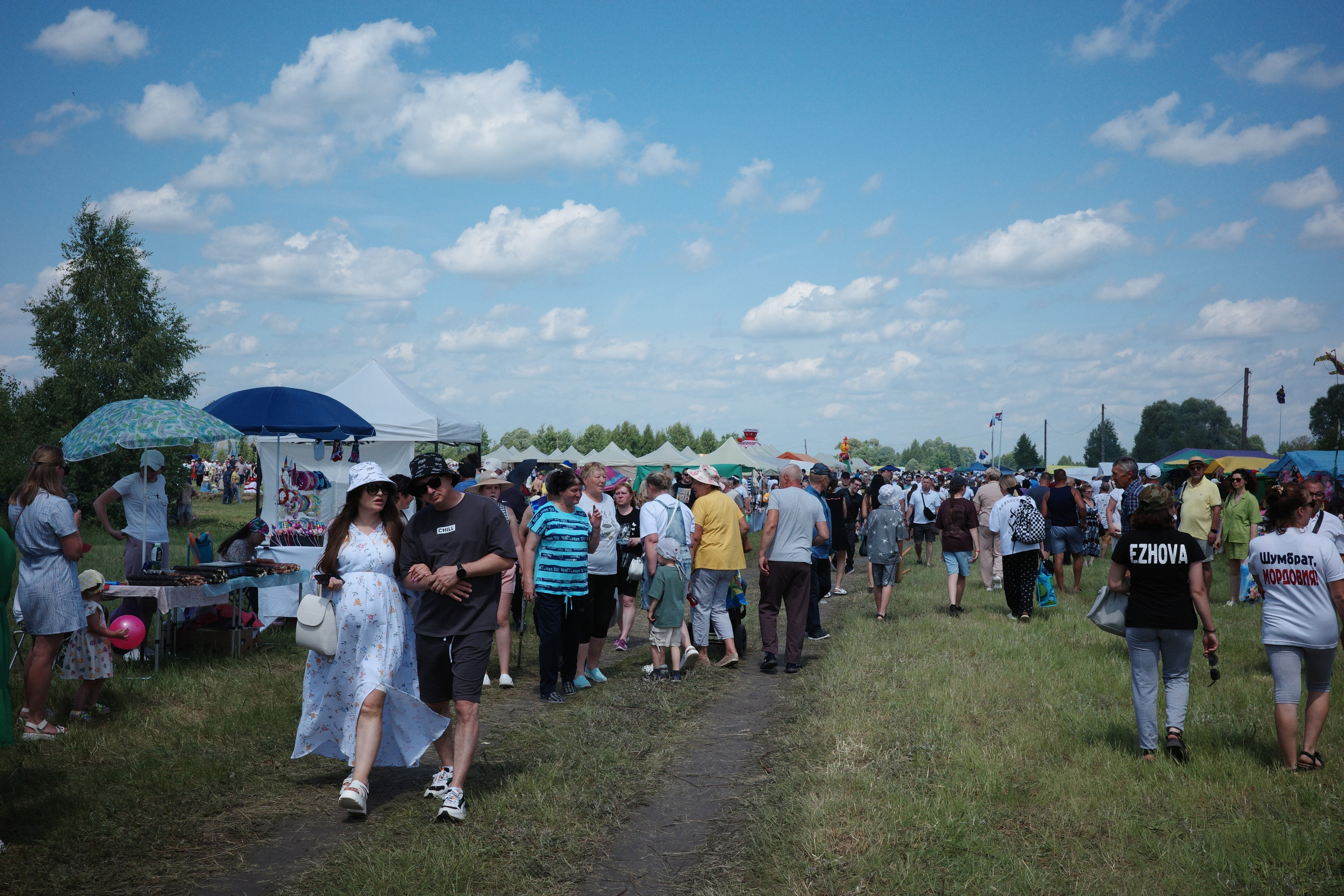 Огромная толпа людей на поле среди белых шатров в солнечный день, голубое небо с облаками