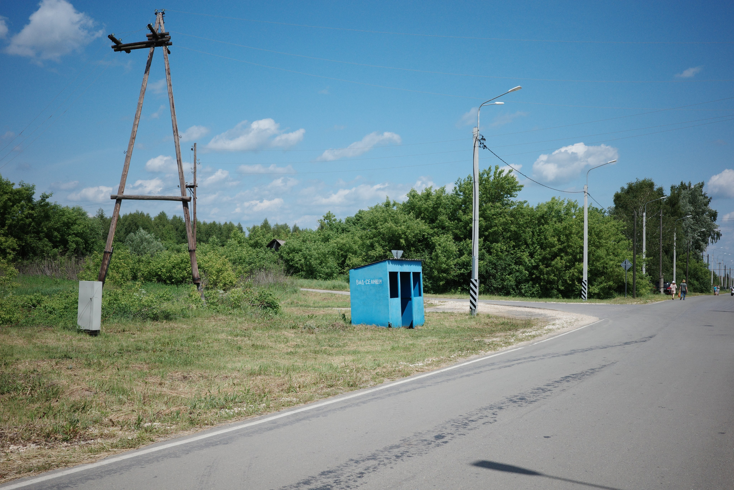 Пустая летняя дорога с синей будкой на обочине, деревья вдали