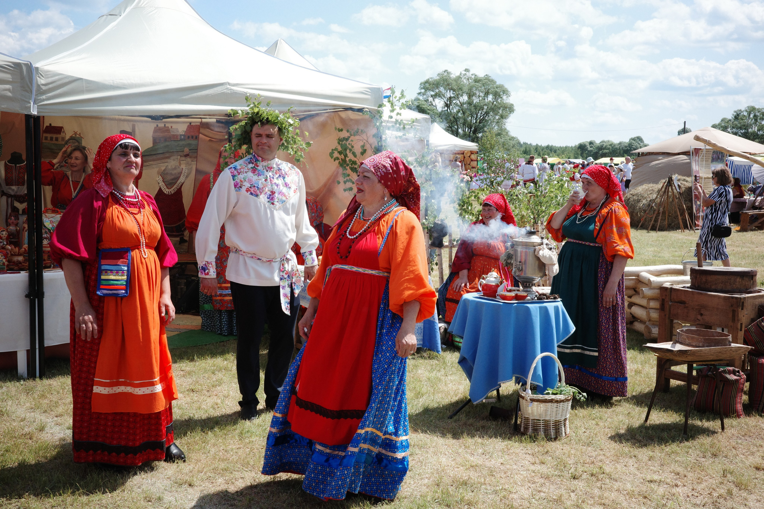 Люди в ярких оранжевых и красных народных костюмах у стенда с самоваром и угощениями
